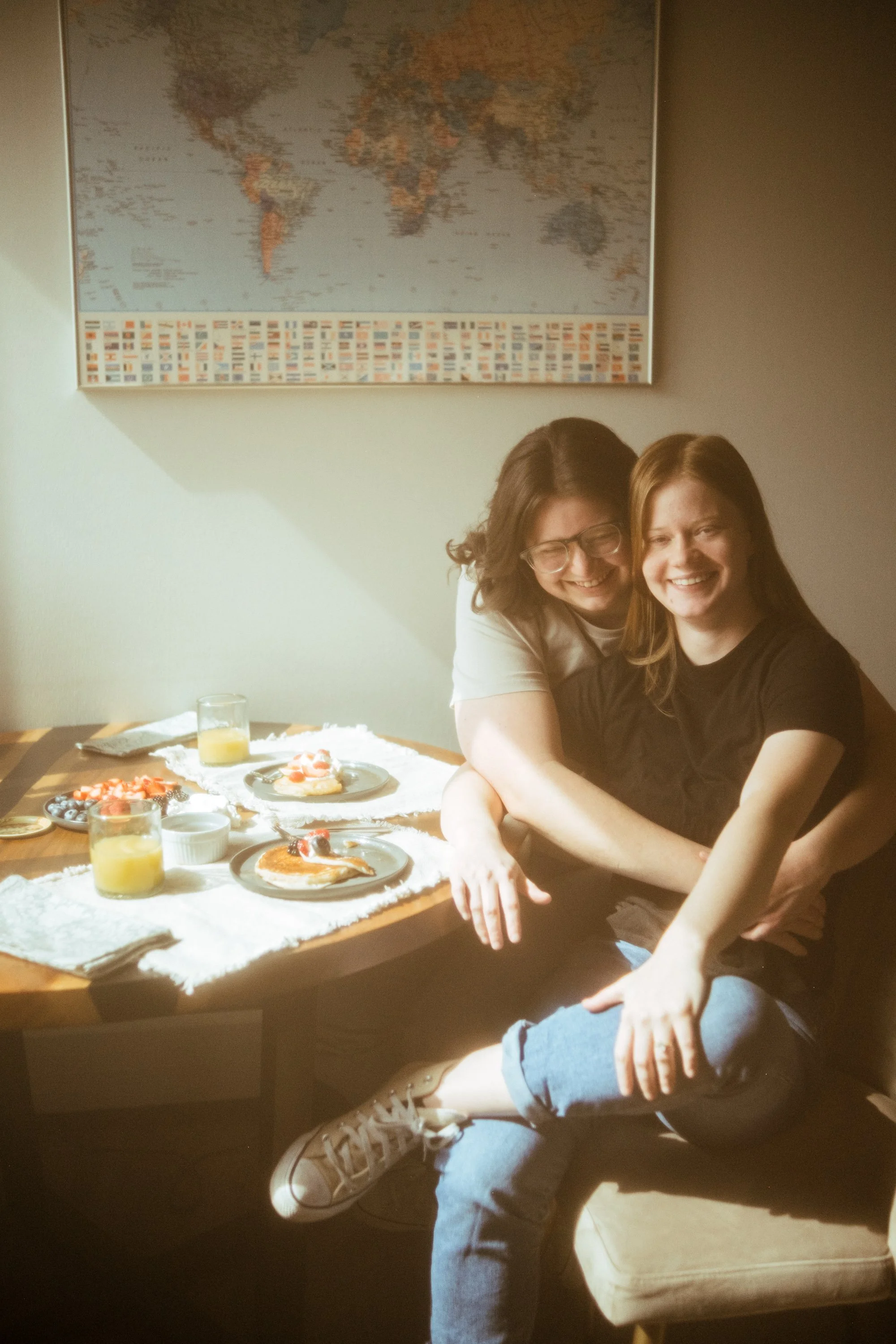 Two women sharing a joyful moment at a breakfast table with pancakes, fruit, and juice, sitting in a room with a world map on the wall.