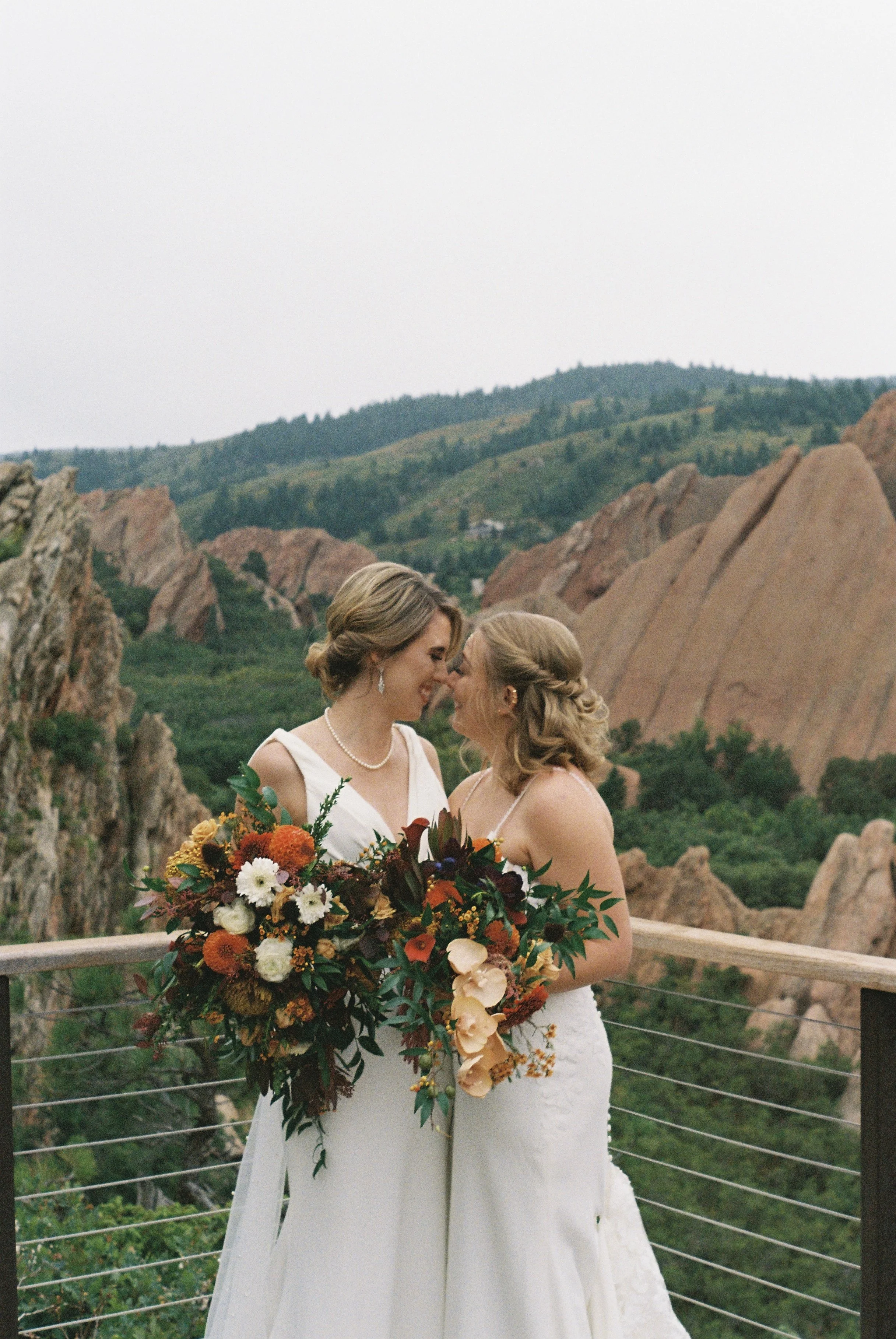 Two women in wedding dresses smiling and leaning close together, holding large colorful bouquets, on a scenic outdoor balcony with rocky hills and green mountains in the background.