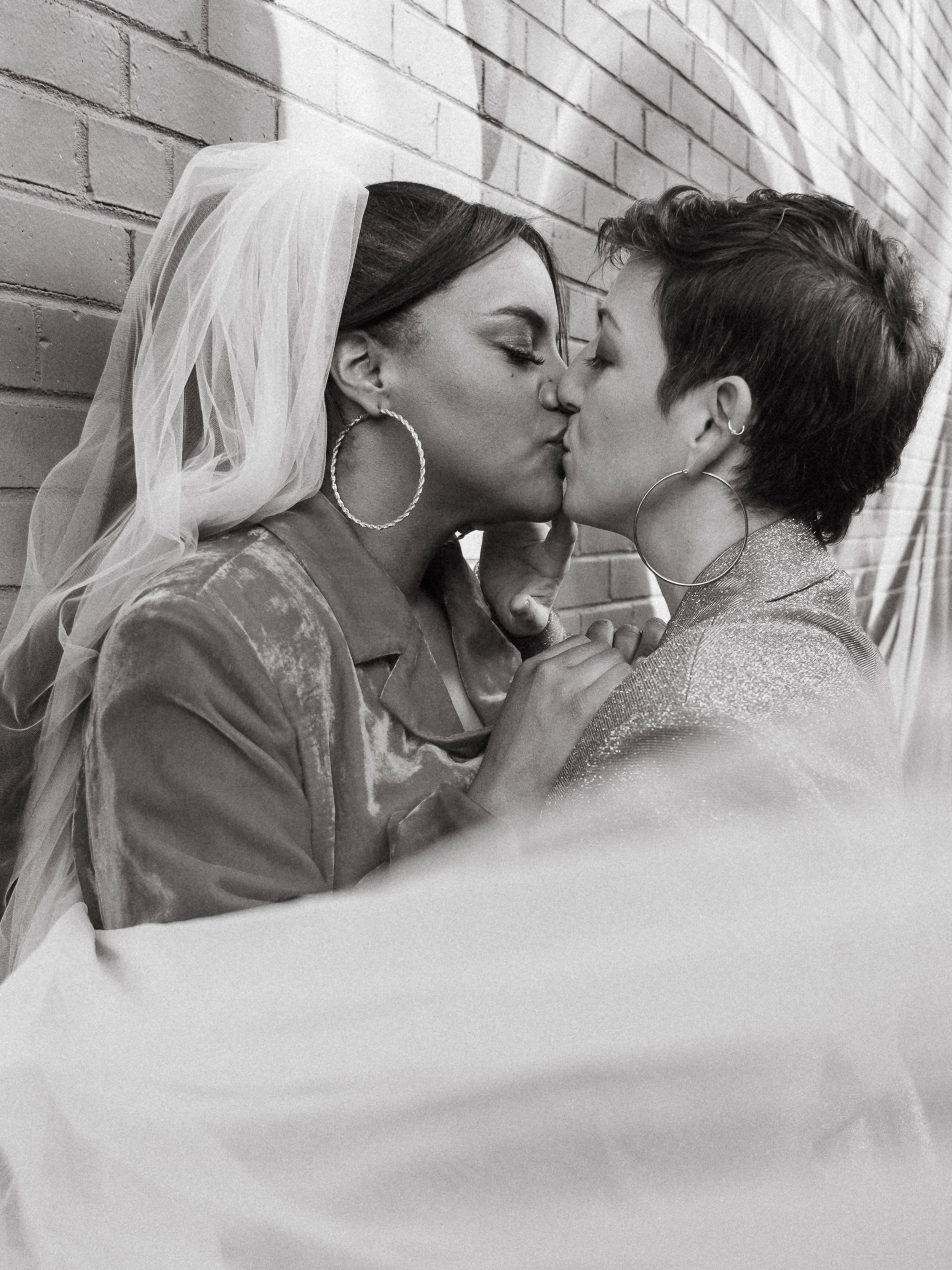 Two women are kissing against a brick wall, one wearing a wedding veil and the other with short hair, both with hoop earrings.