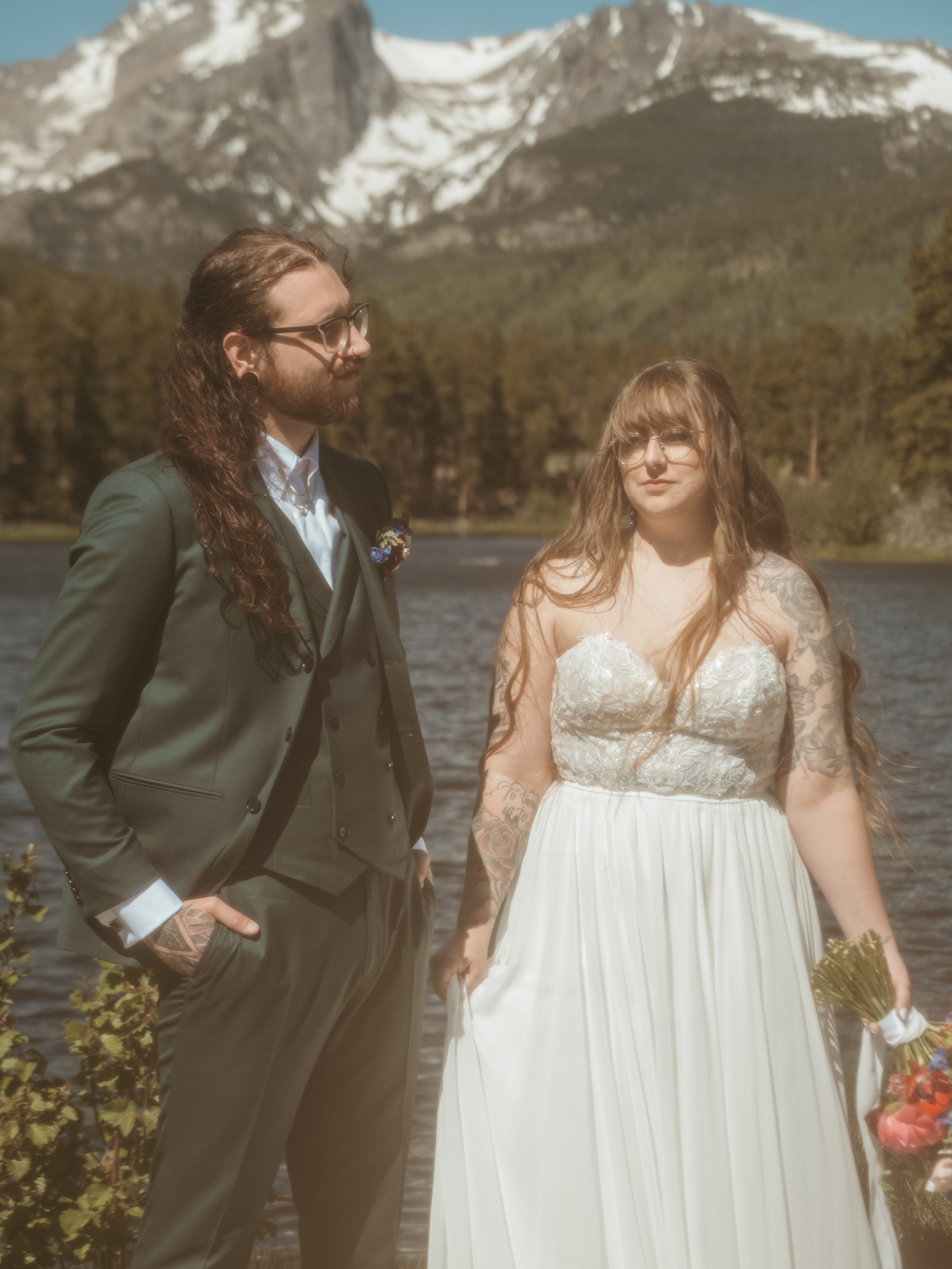 A couple dressed in wedding attire standing beside a lake with snow-capped mountains and trees in the background.