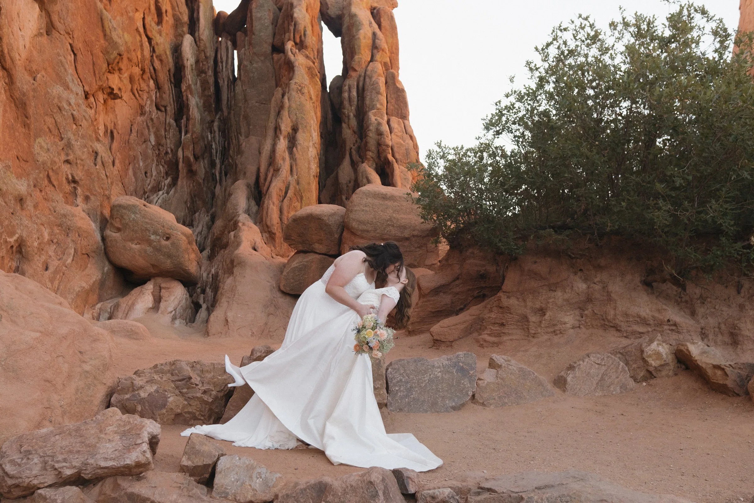 Two women in white wedding dresses sharing a kiss in a rocky desert landscape with red sandstone formations and a green shrub.