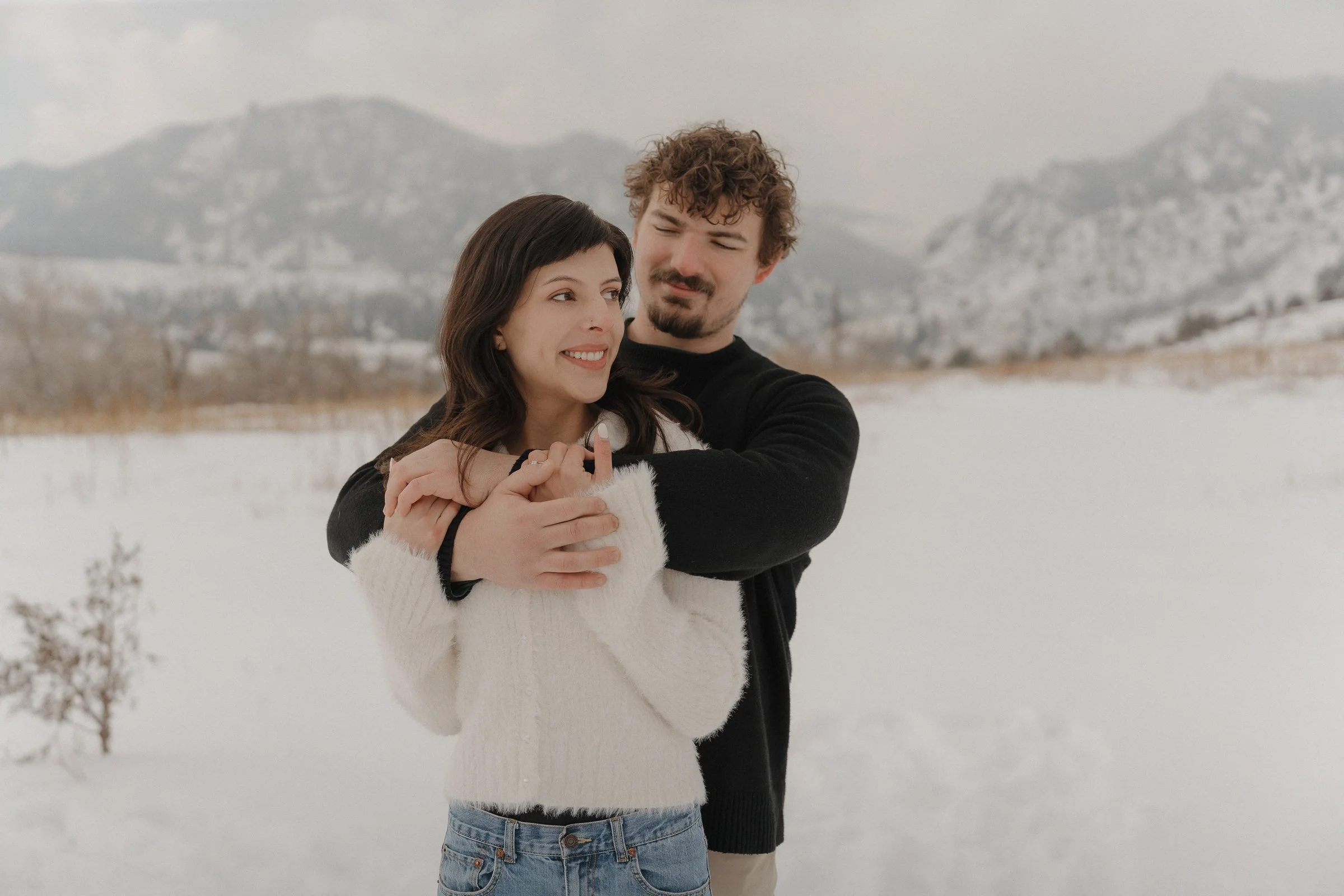 A couple embraces in a snowy landscape with mountains in the background, the woman wearing a white sweater and the man in a black sweater.