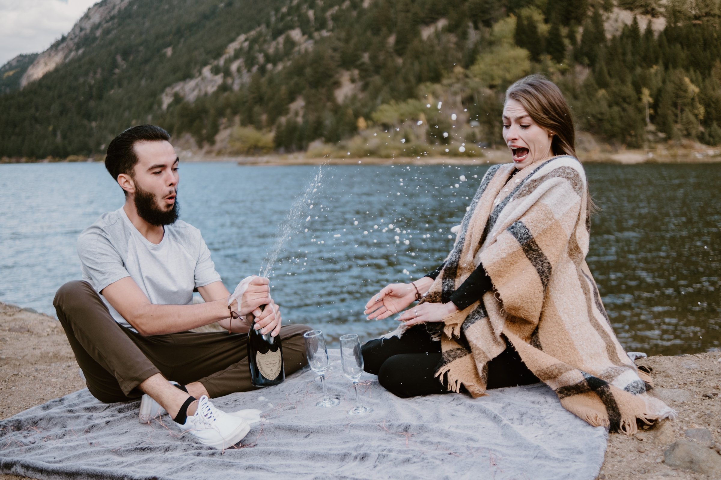 A man and woman having a picnic by a lake, with the man opening a champagne bottle causing it to spray, and the woman reacting surprisedly, all sitting on a blanket with champagne glasses nearby, surrounded by mountains and trees.