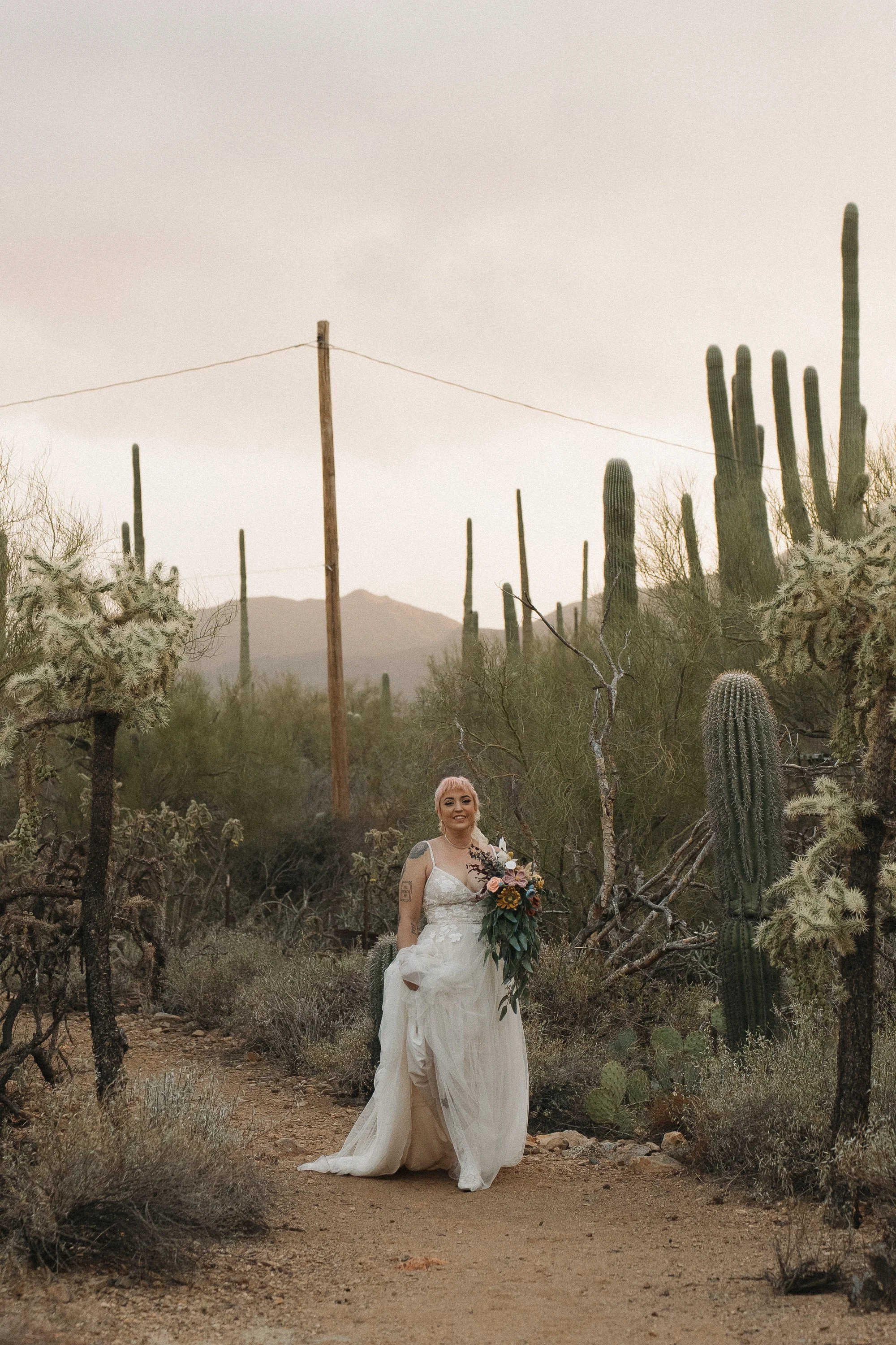 A smiling woman in a white wedding dress holding a bouquet of flowers walking along a desert trail surrounded by cacti and desert shrubs, with mountains in the background.