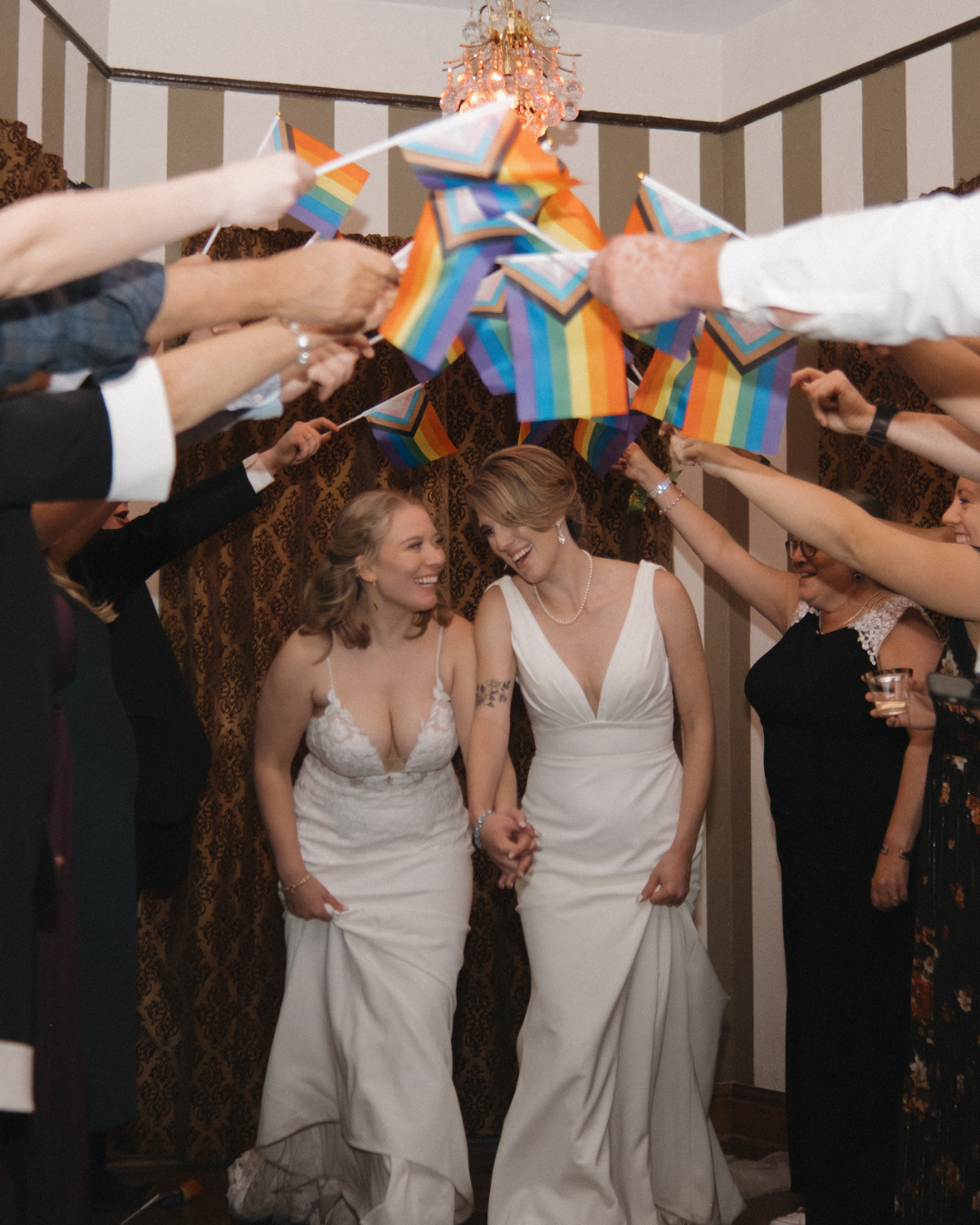 Two women in white wedding dresses hold hands and smile at each other in the middle of a rainbow flag banner held by surrounding people, celebrating at a wedding.