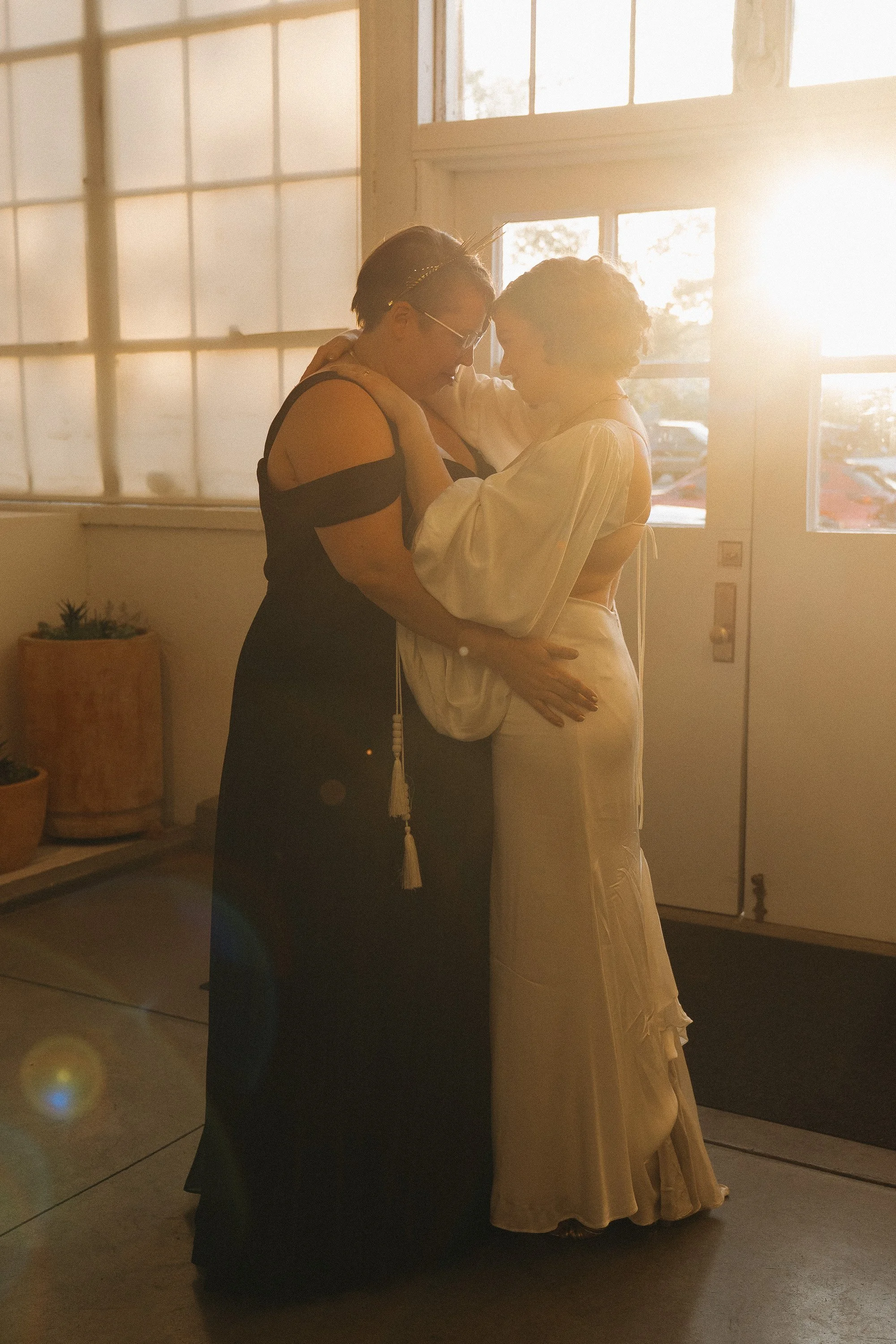 Two women dancing closely together in a warmly lit room with large windows and sunlight streaming in.