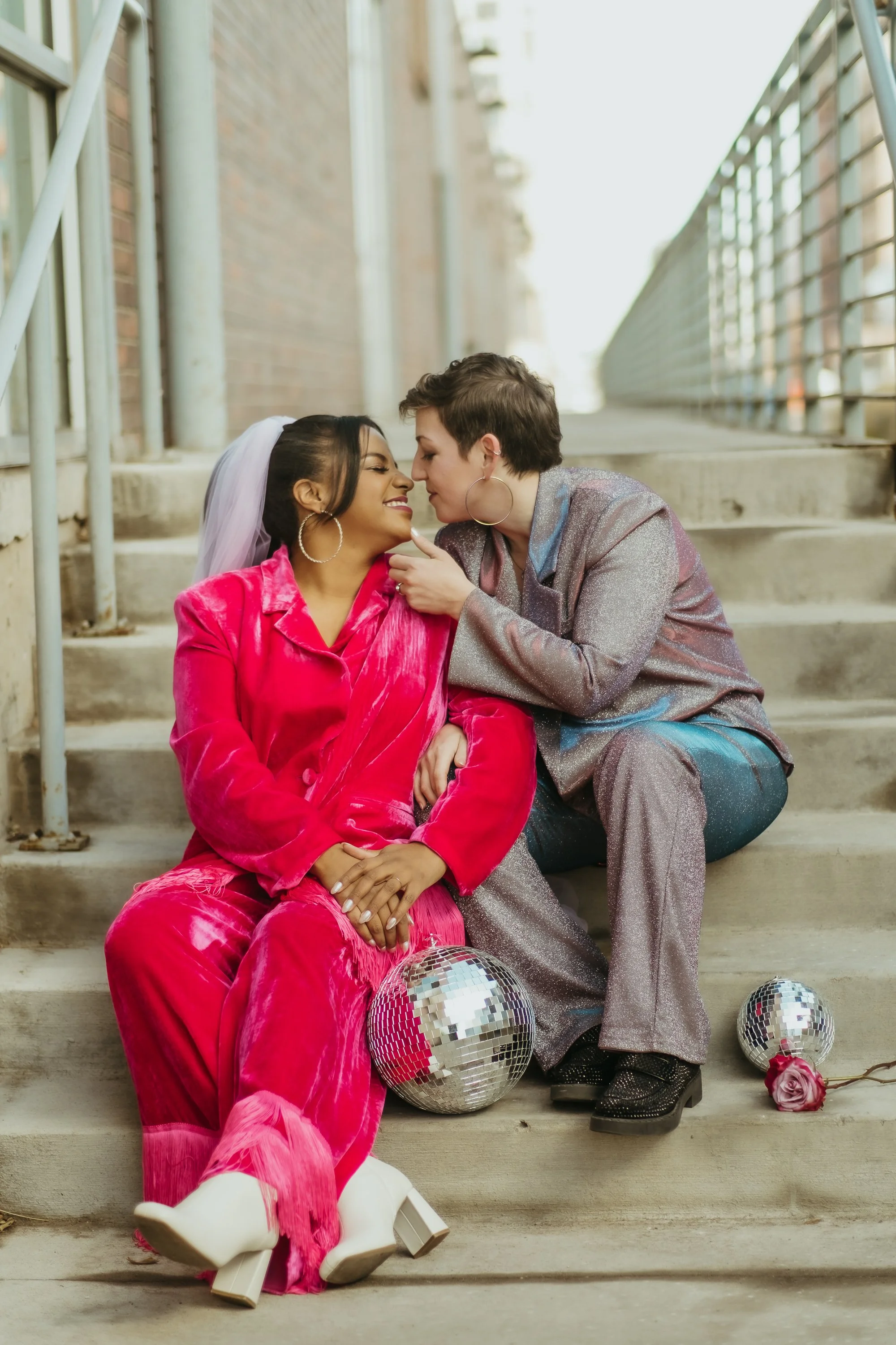 Two women with short hair and hoop earrings sit on outdoor stairs, leaning close face to face, sharing an intimate moment. One wears a bright pink velvet suit with a veil, the other wears a shiny silver suit. Disco balls and a pink rose are next to t