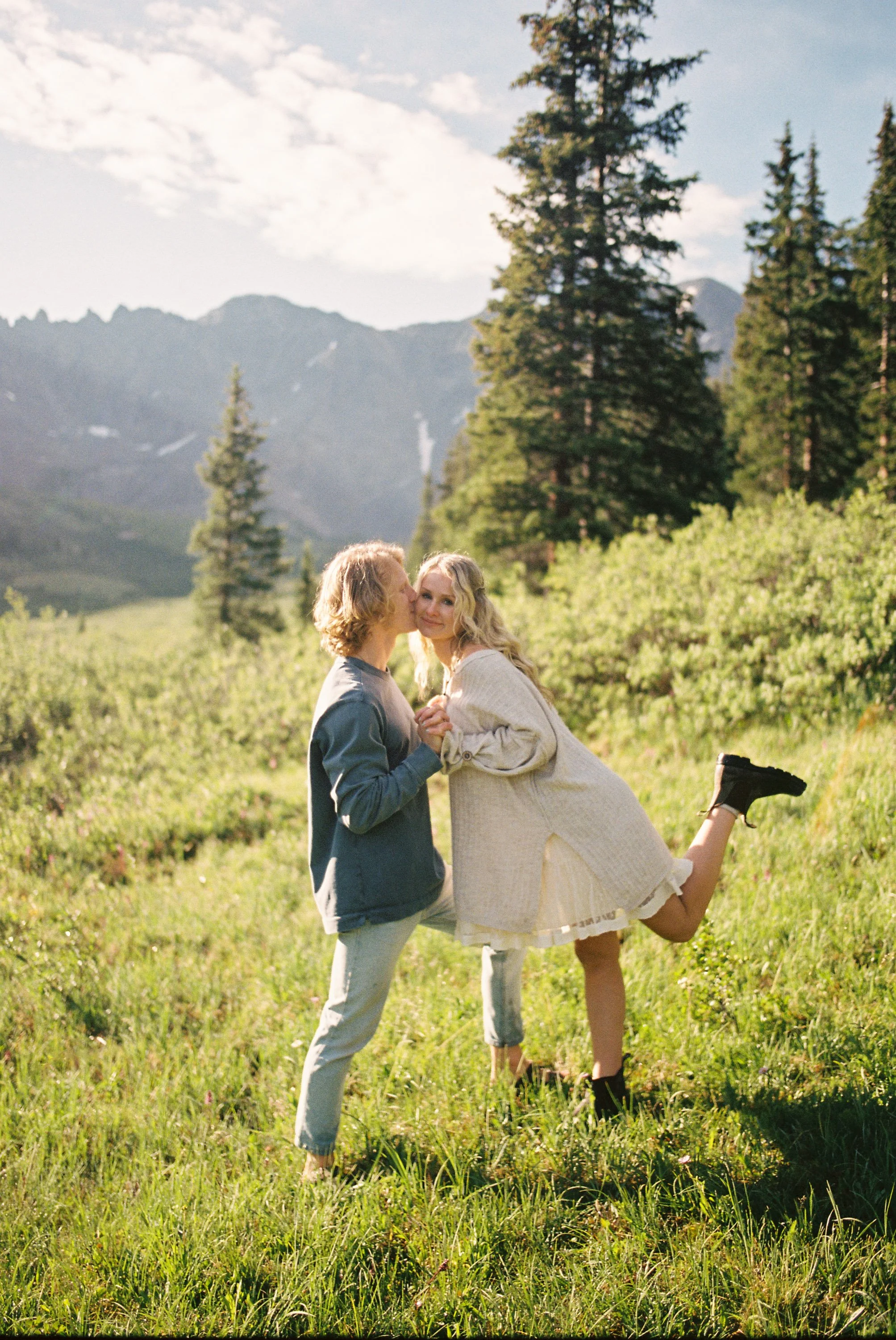 A couple standing in a grassy field with mountains and pine trees in the background, the man kissing the woman on the cheek, the woman smiling and lifting one leg.