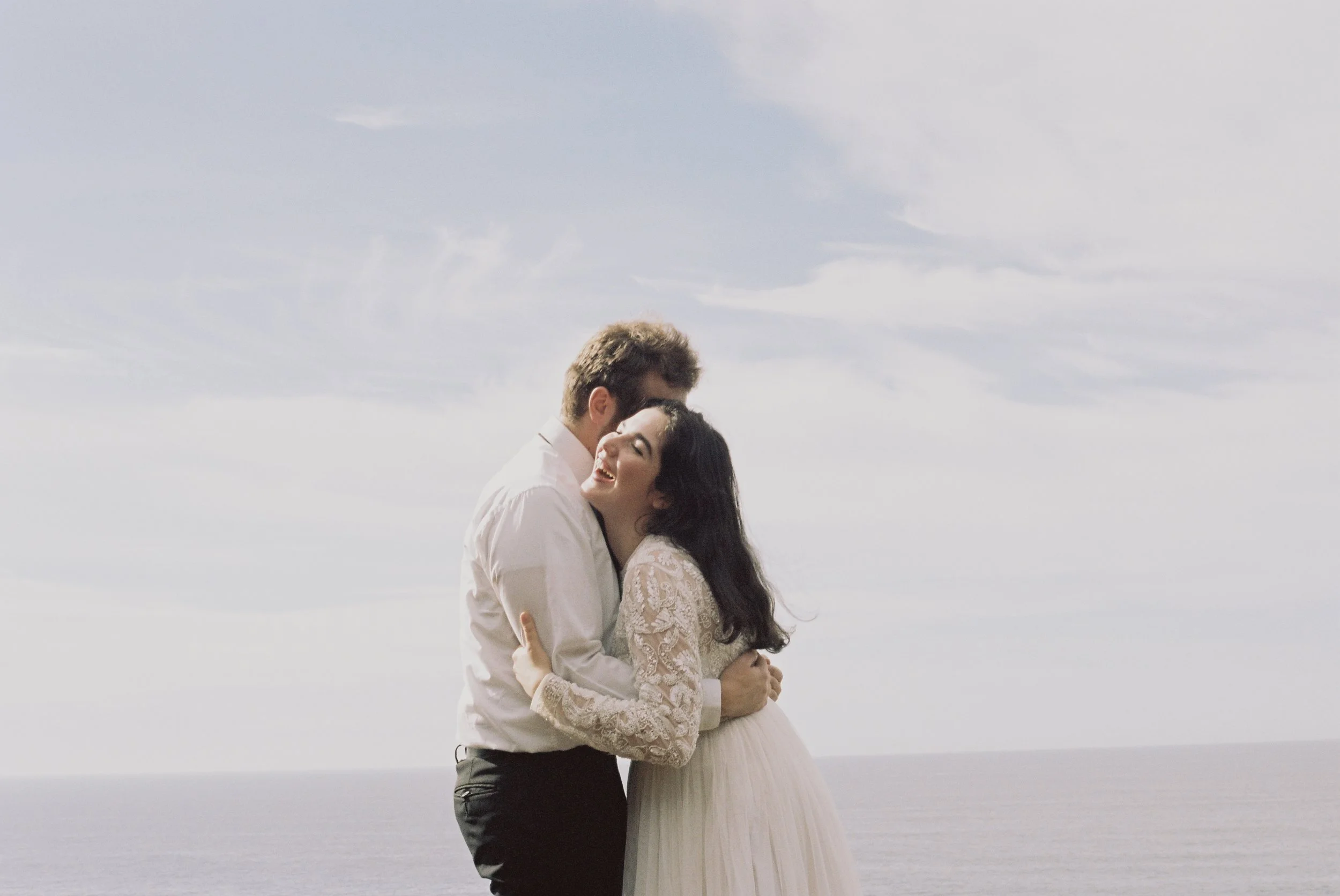 A couple hugging by the ocean, smiling and enjoying the moment, with a cloudy sky in the background.