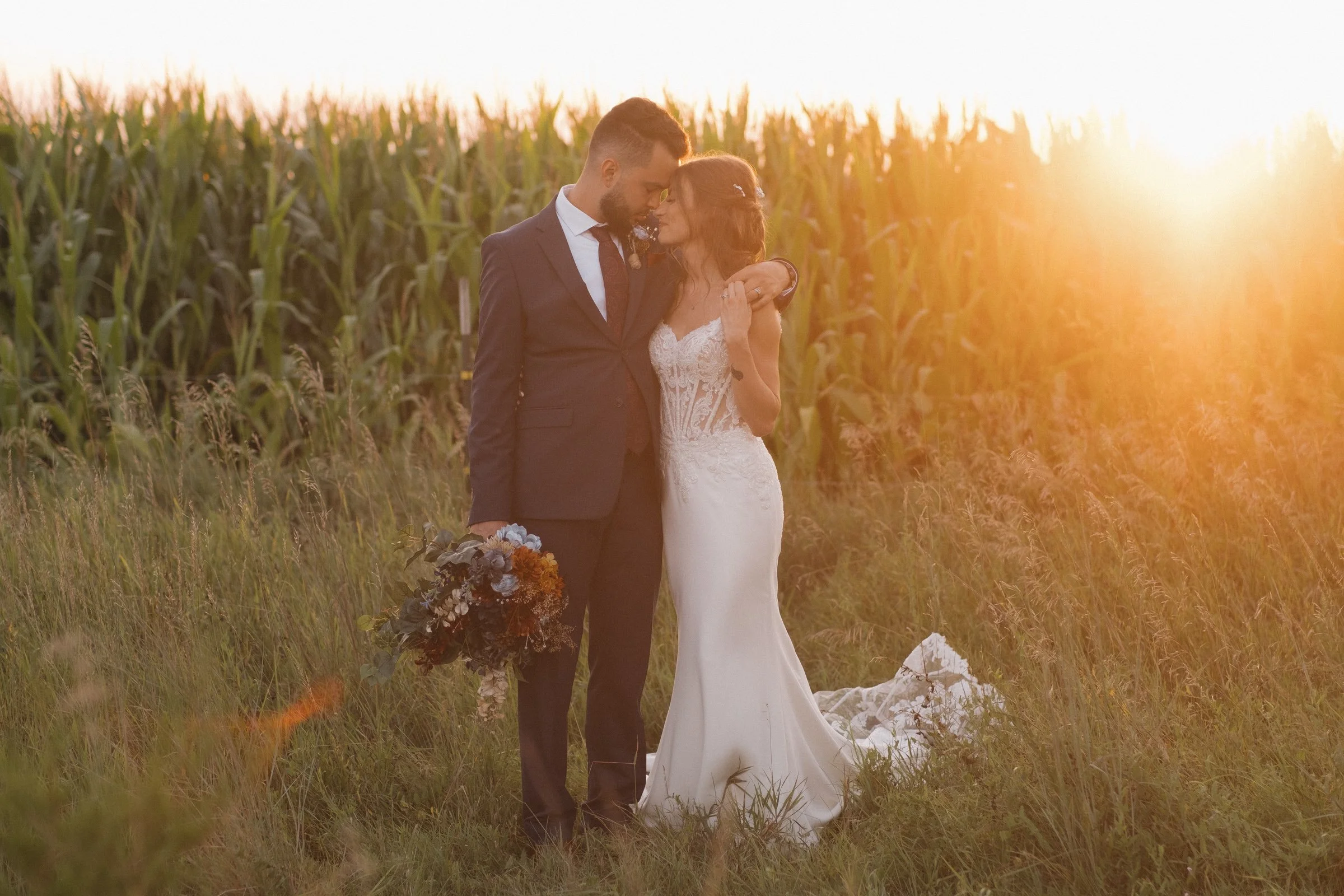 A newlywed couple stands closely together in a field of grass and tall plants, embracing each other during sunset. The groom wears a dark suit and holds a bouquet, while the bride is in a white lace wedding dress. The sky is glowing with warm sunligh