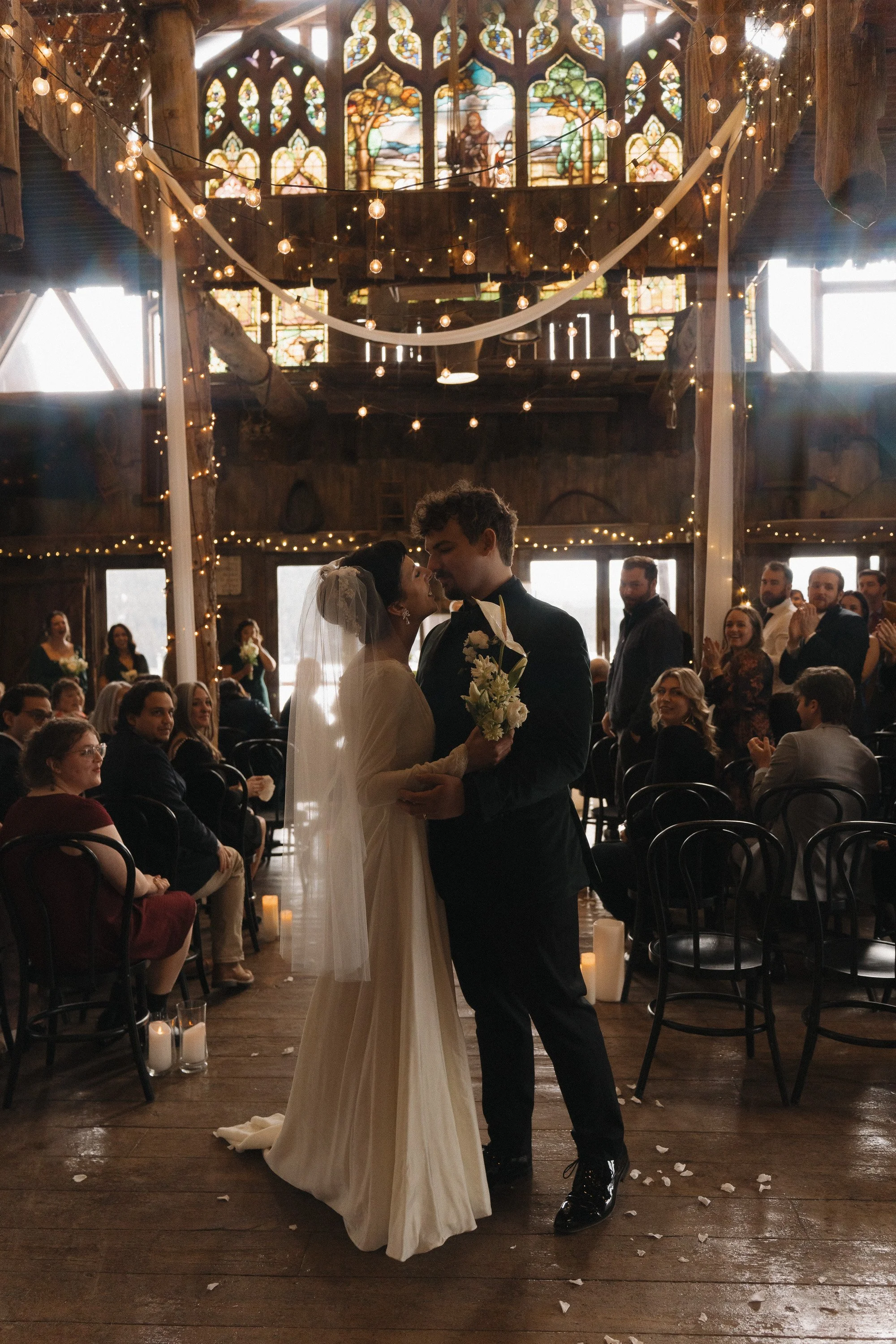 Bride and groom sharing a dance during their wedding ceremony in a warmly decorated rustic venue with stained glass windows, string lights, and surrounded by seated guests.