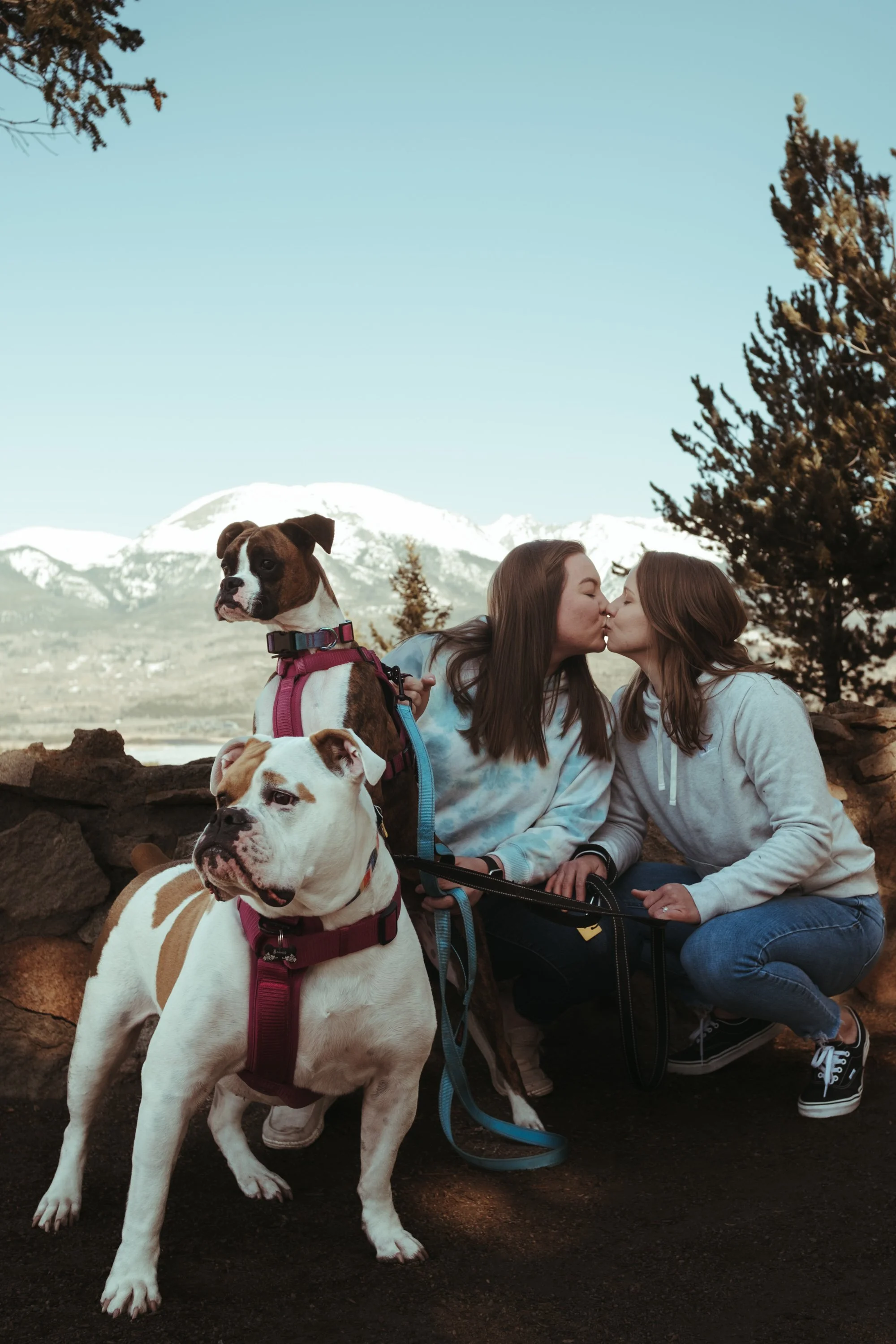 Two women share a kiss outdoors with two dogs, mountains, and trees in the background.