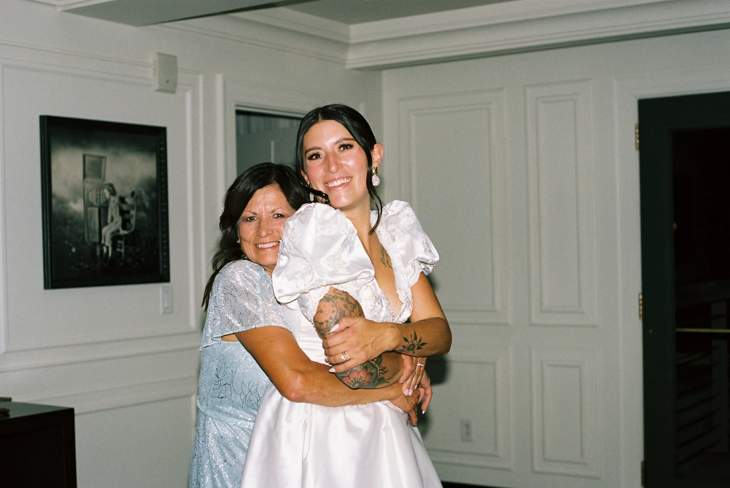 Two women, a bride in a white dress and a woman in a silver dress, hugging and smiling in a well-lit room with white paneling and a framed painting on the wall.