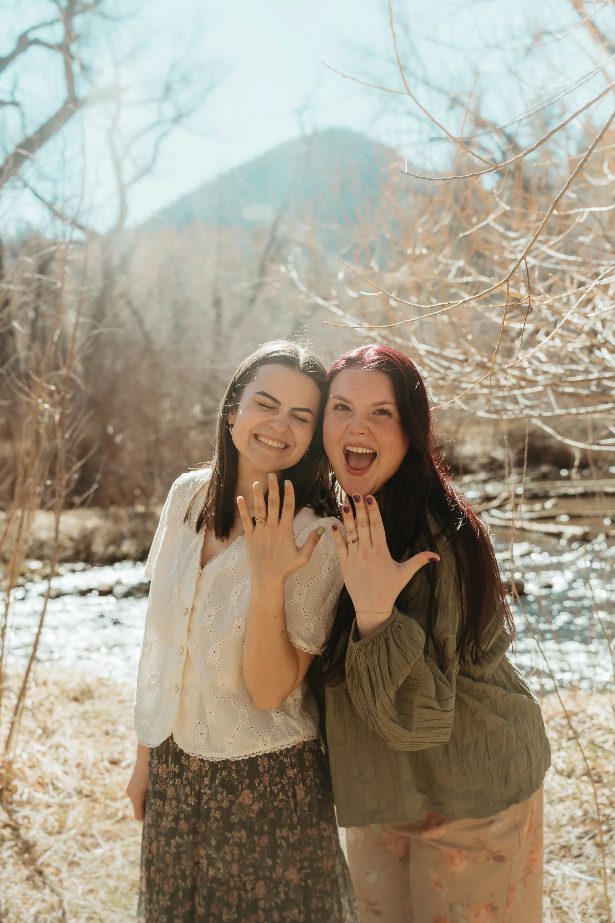 Two women smiling and showing off rings on their fingers outdoors on a sunny day with trees and a mountain in the background.