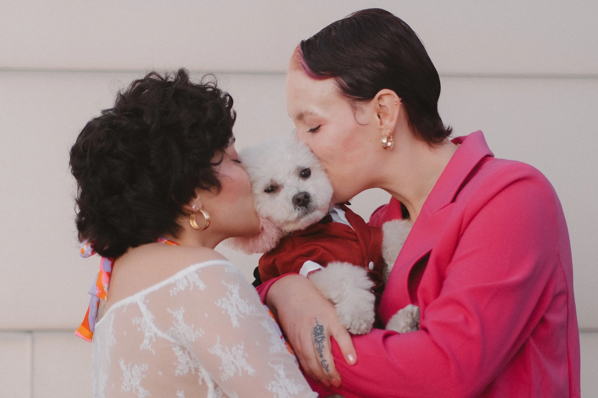 Two women giving kisses to a small white dog dressed in red, holding the dog between them. One woman has short, curly dark hair and is wearing a white lace top with a colorful scarf. The other woman has short dark hair with pink highlights, gold earr