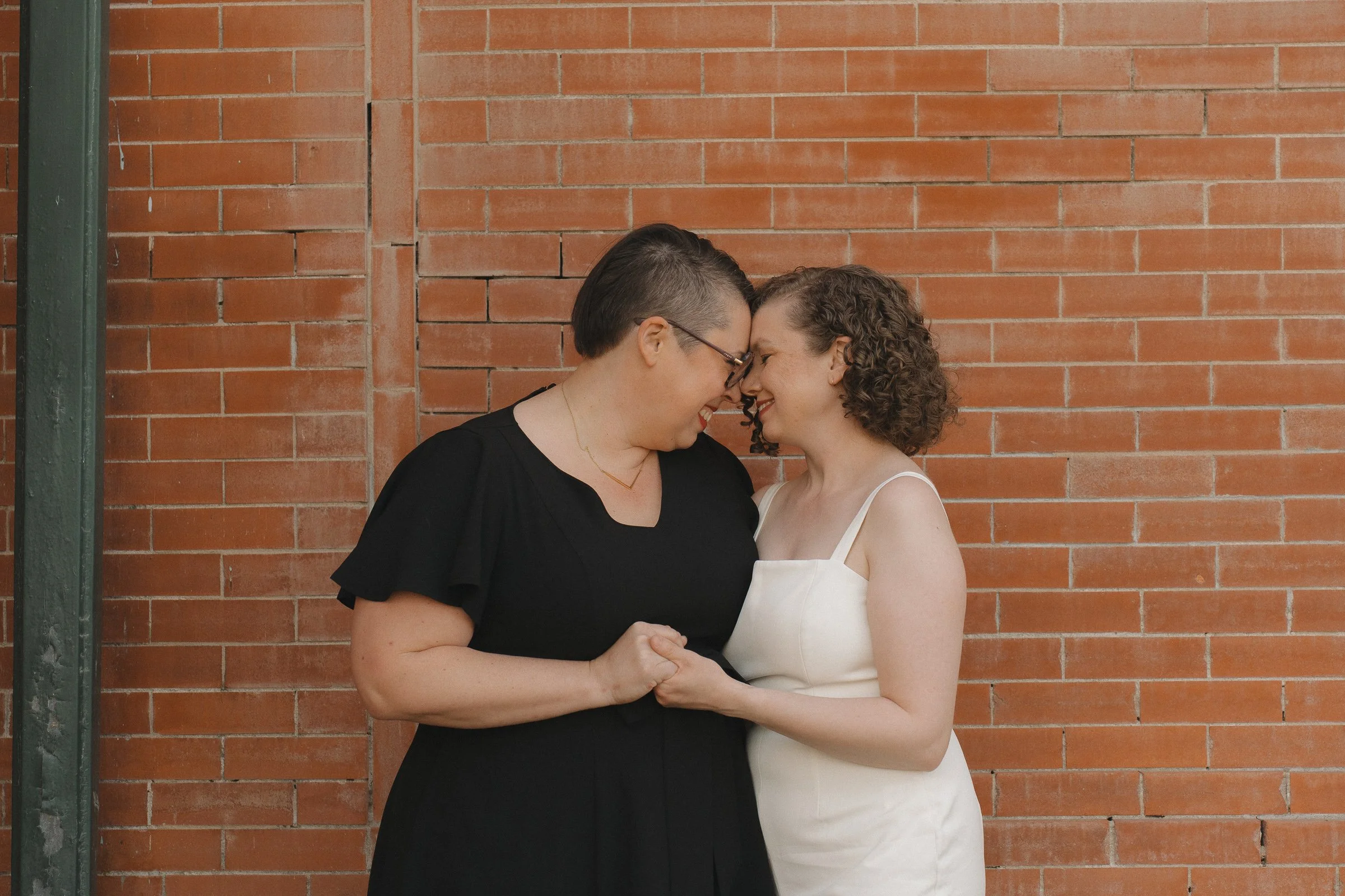 Two women with their foreheads touching, holding hands, smiling, standing against a red brick wall.