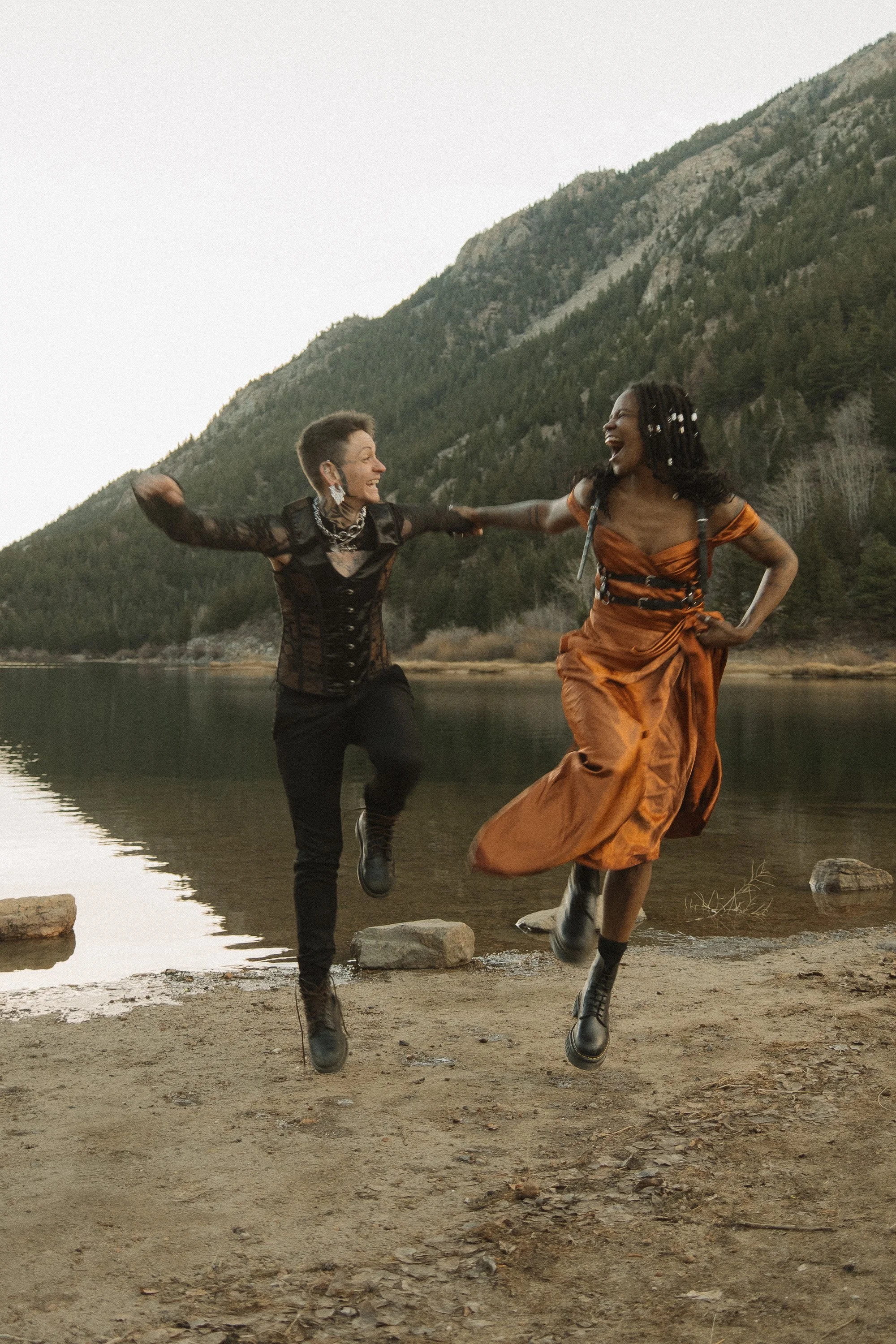 Two women joyfully jumping and holding hands by a lakeshore with forested mountains in the background.