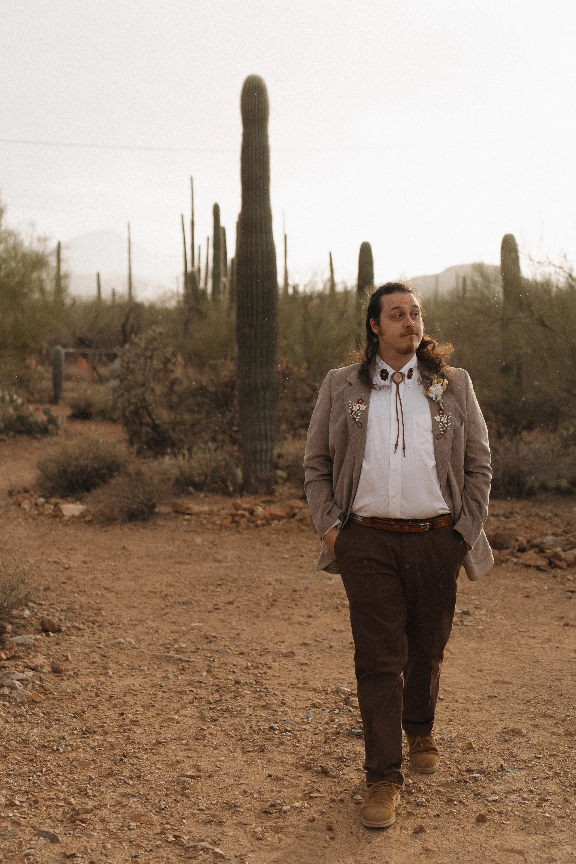A man with long hair walking in a desert landscape with cacti, wearing a beige blazer with floral embroidery, a white shirt, dark trousers, and tan boots.
