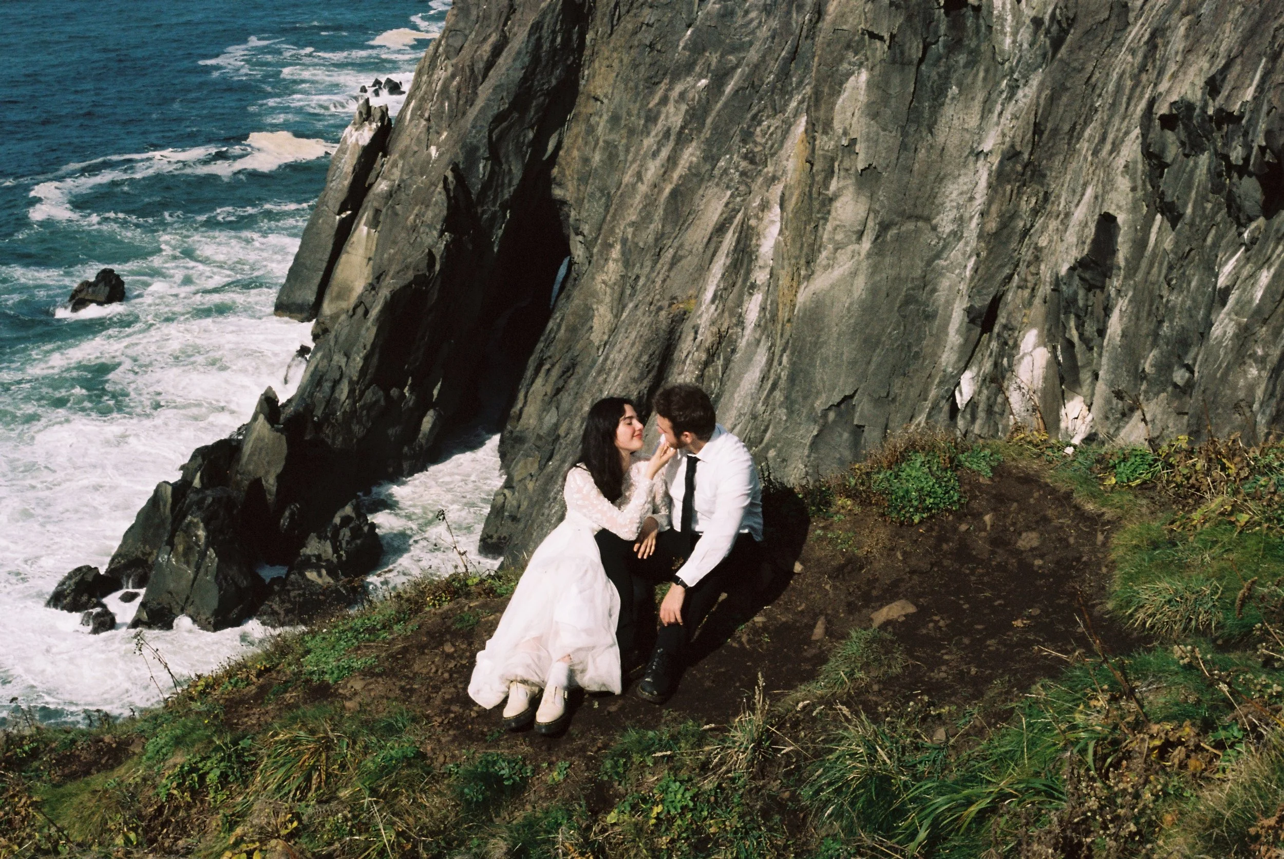 A young couple sitting on a grassy hillside near the ocean, with large rocky cliffs in the background. The woman wears a white dress and the man a white shirt with a black tie. They are close to each other, gazing into each other's eyes.