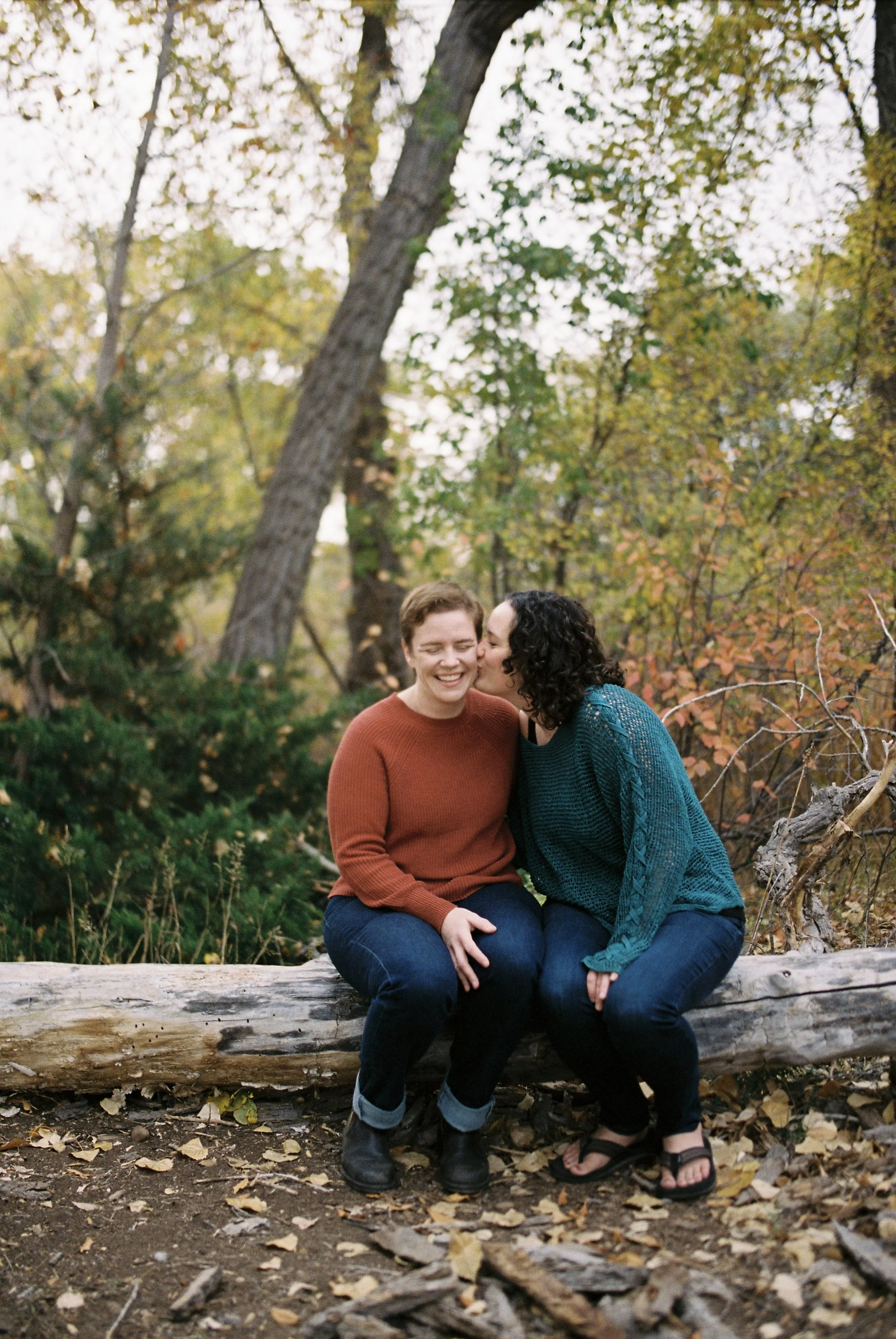Two women sitting on a fallen log outdoors, one whispering into the other's ear, surrounded by autumn foliage.