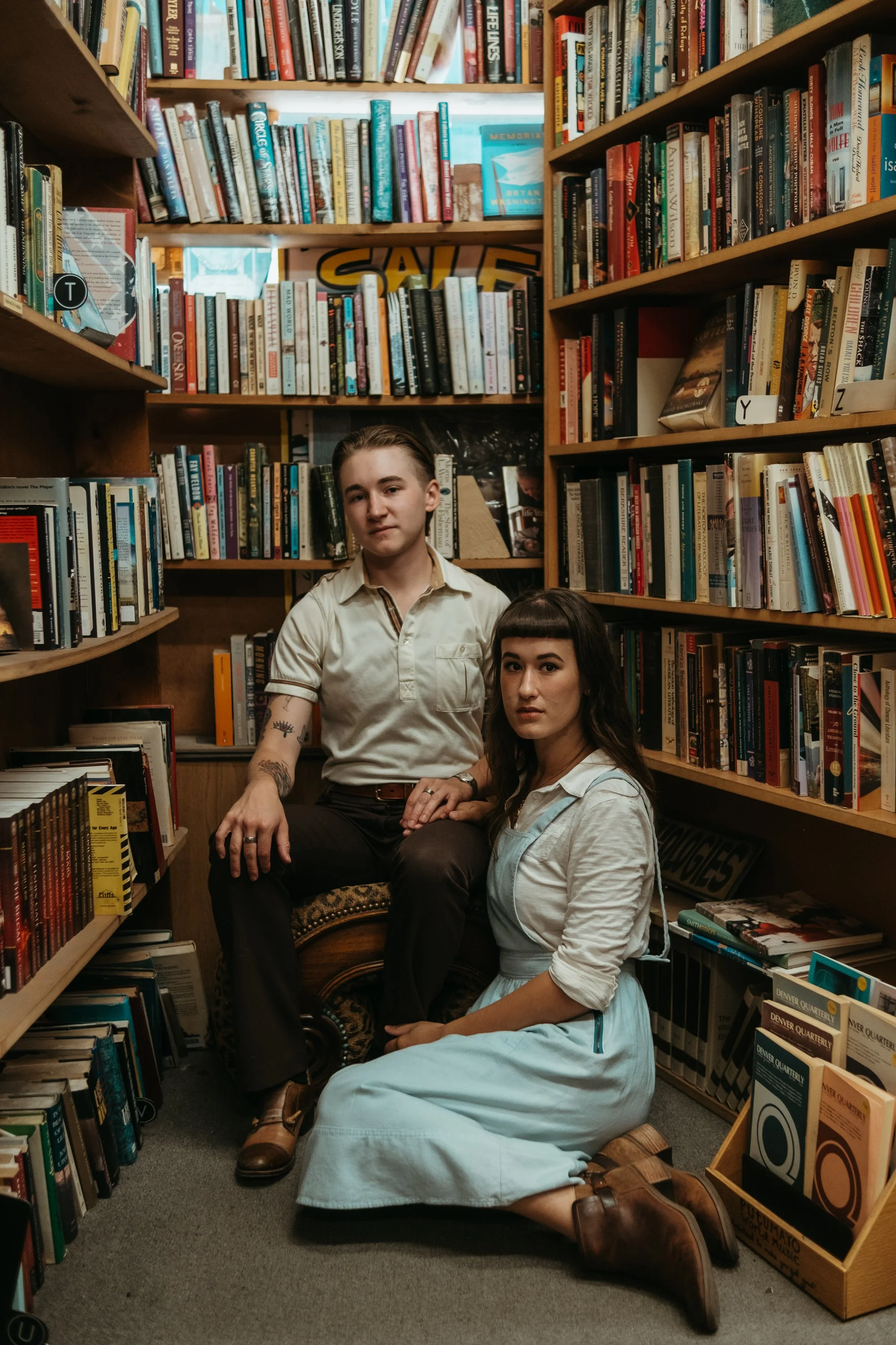 Two women in vintage clothing posing in a small bookstore aisle with bookshelves filled with magazines and books around them.