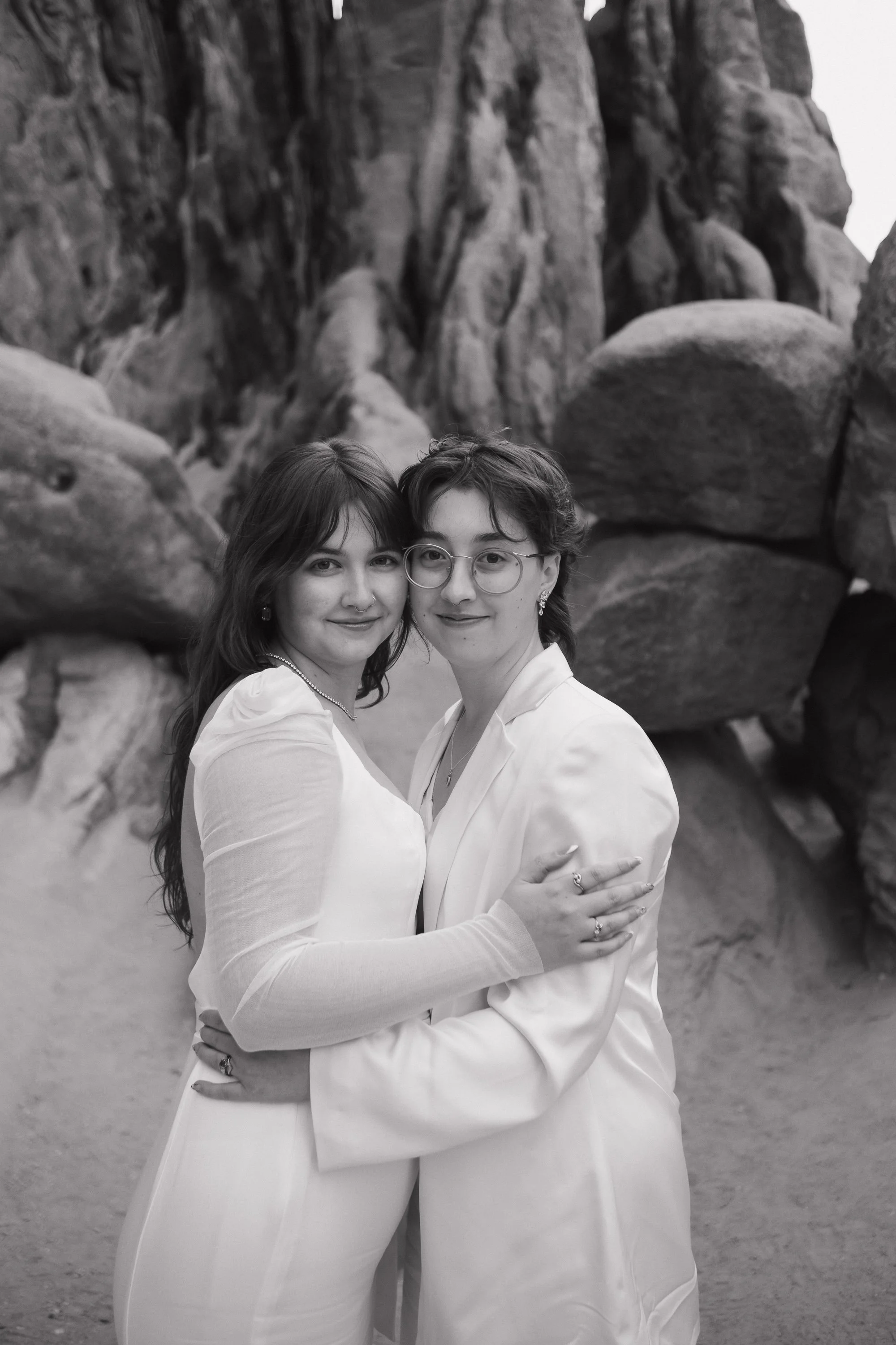 Two women embracing in front of rocky cliffs, both dressed in white outfits, smiling at the camera.