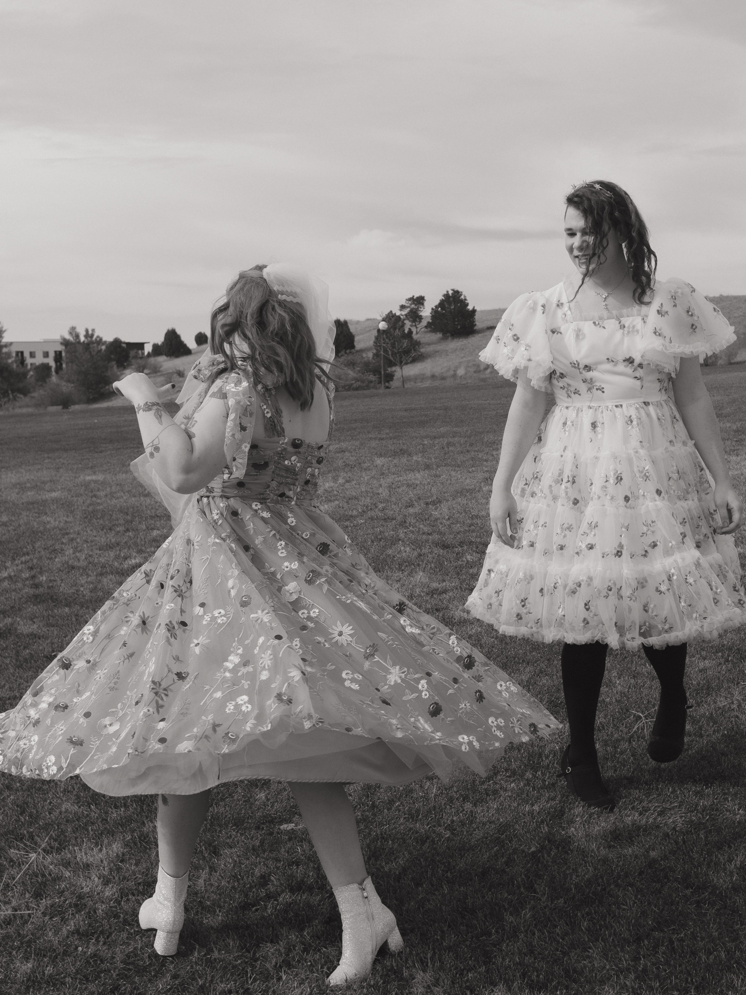 Two women in vintage-style floral dresses dancing in an open grassy field with a cloudy sky.