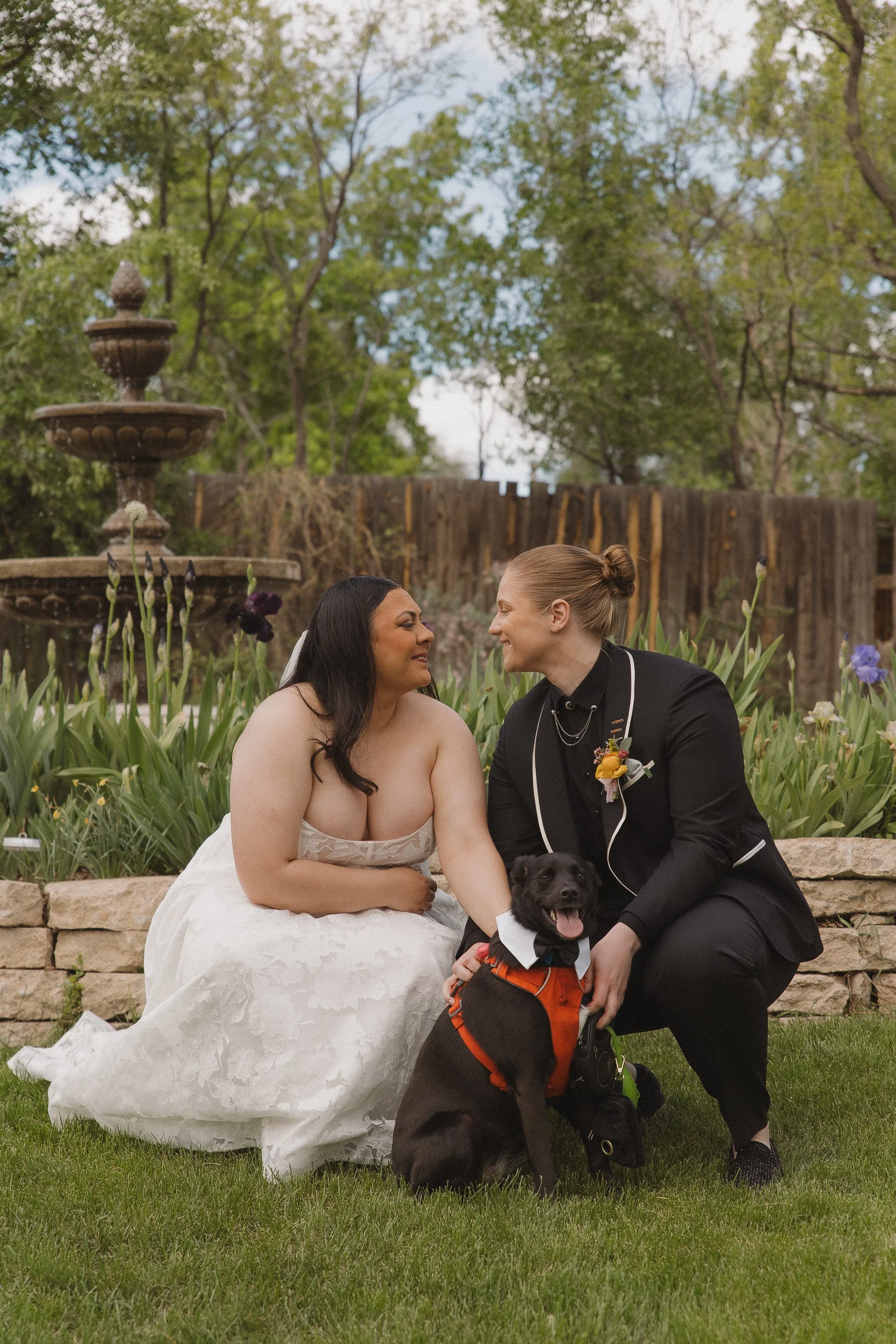 A same-sex couple in wedding attire sitting outdoors with their service dog, smiling and looking into each other's eyes. The bride in a white wedding gown and the other in a black suit, with a garden and fountain in the background.