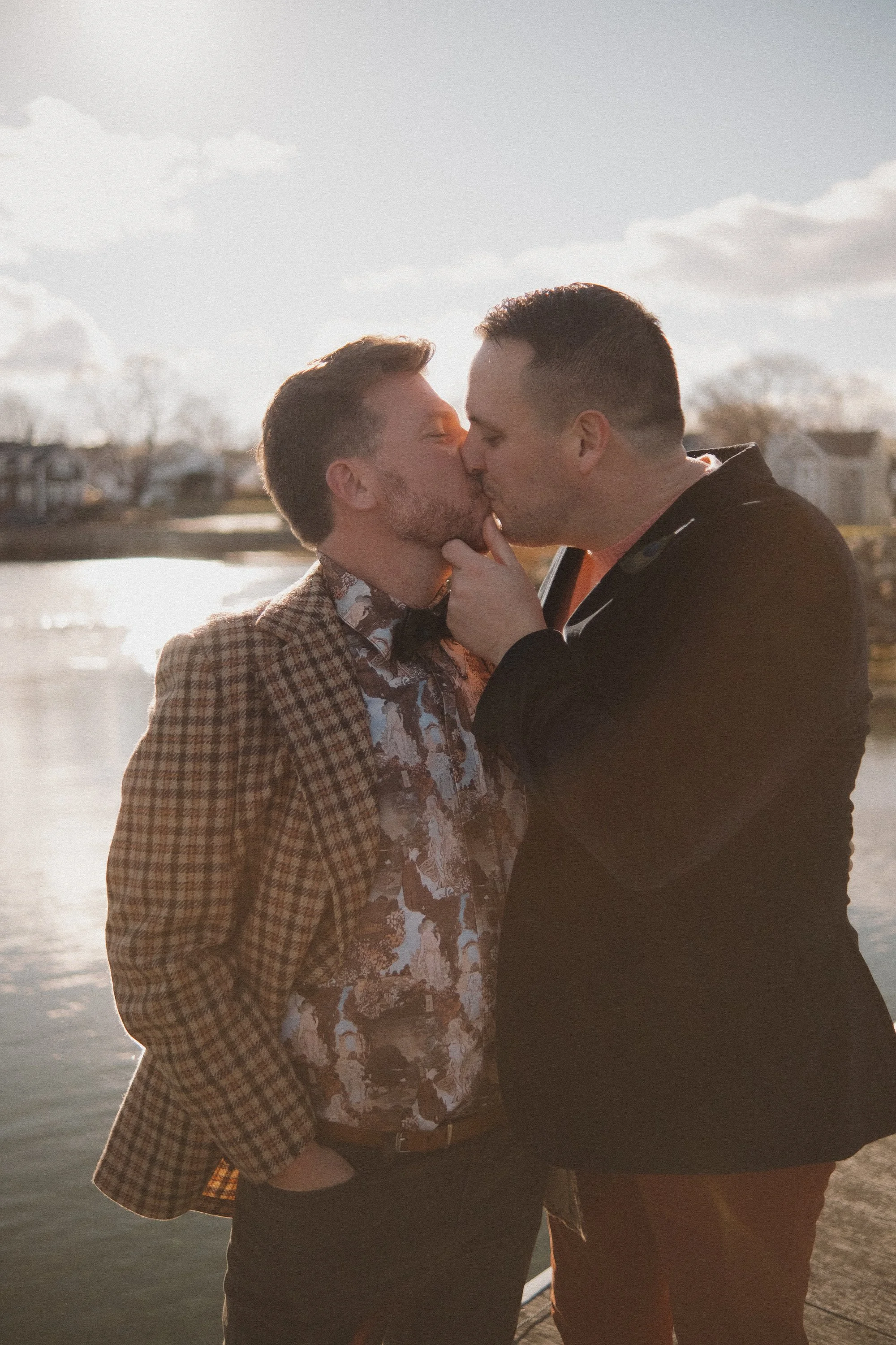 Two men kissing outdoors near a body of water with houses in the background.