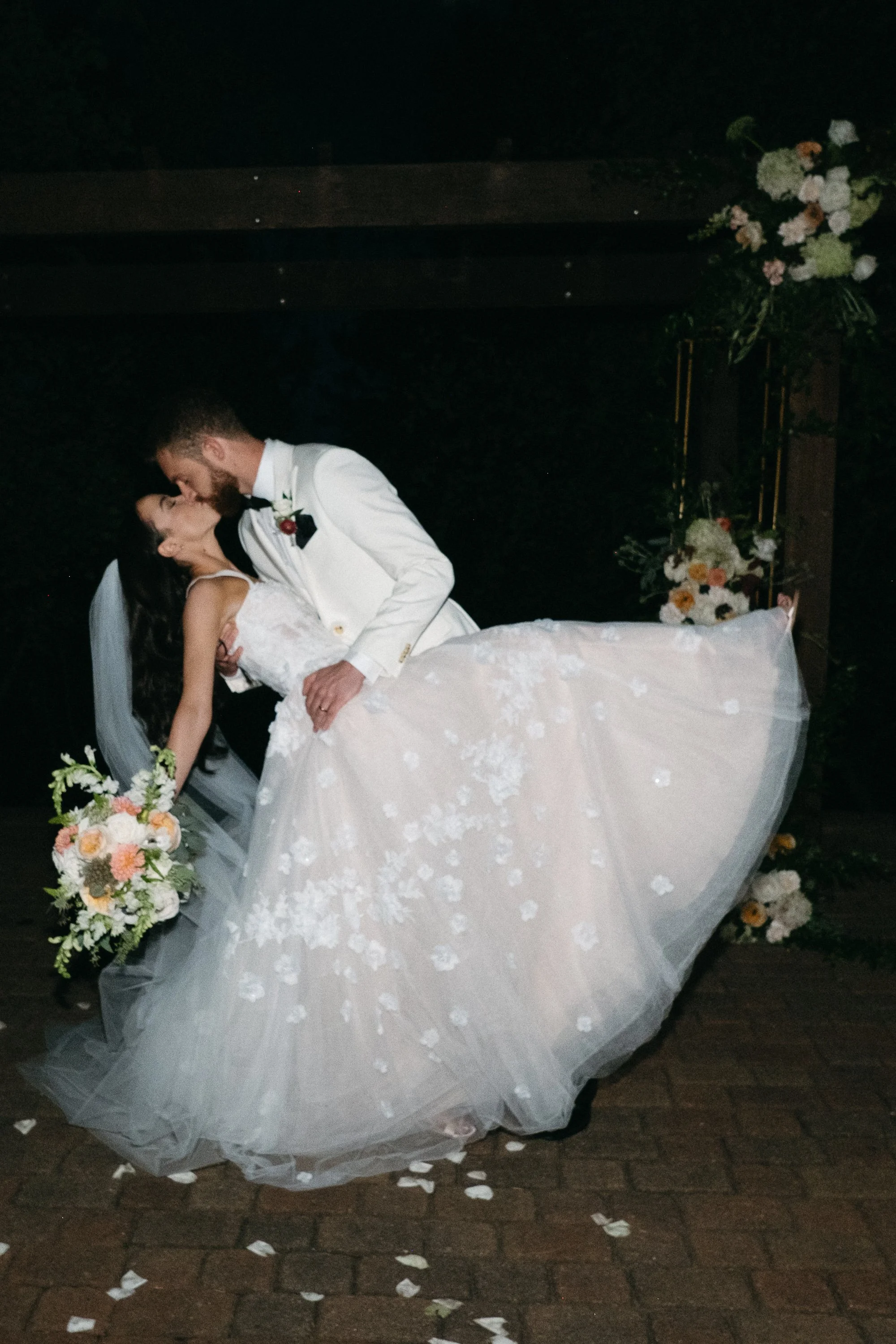 A bride and groom sharing a kiss at their wedding, with the groom holding the bride while she leans back. The bride is wearing a white wedding gown and holding a bouquet. The scene is decorated with flowers.