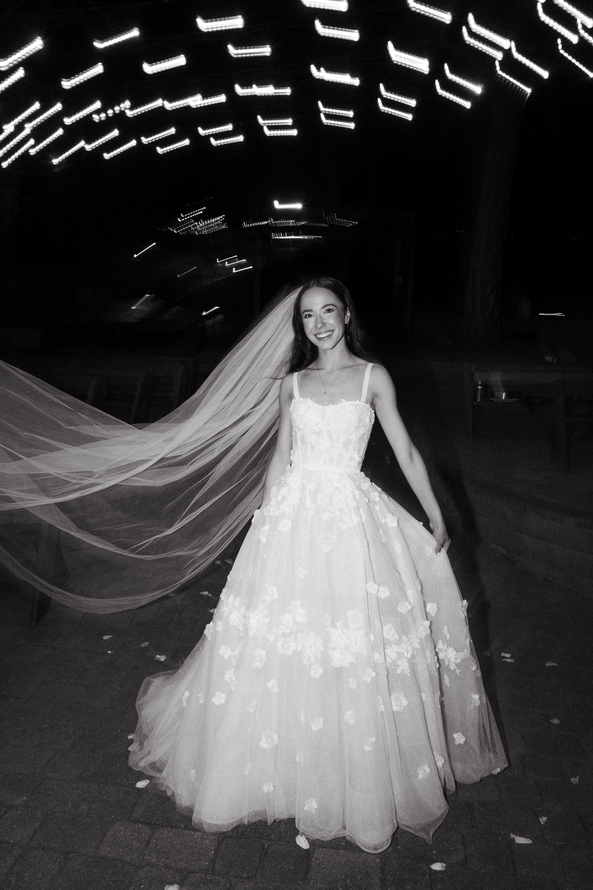 Black and white photo of a smiling bride wearing a wedding gown with floral details and a veil, standing on a dark outdoor area with blurred lights overhead.