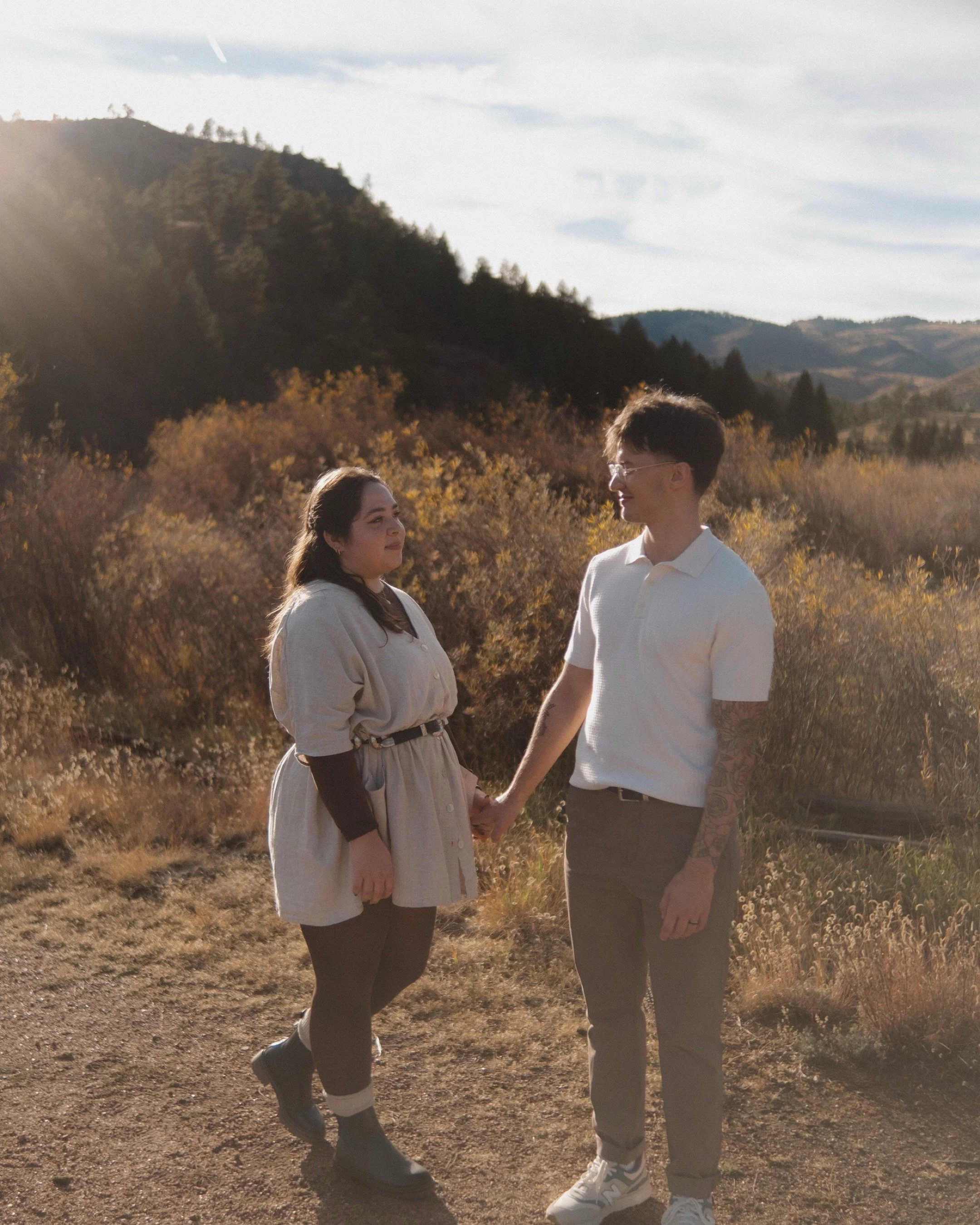 A young couple holding hands in a scenic outdoor setting during sunset, with mountainous terrain and autumn foliage in the background.