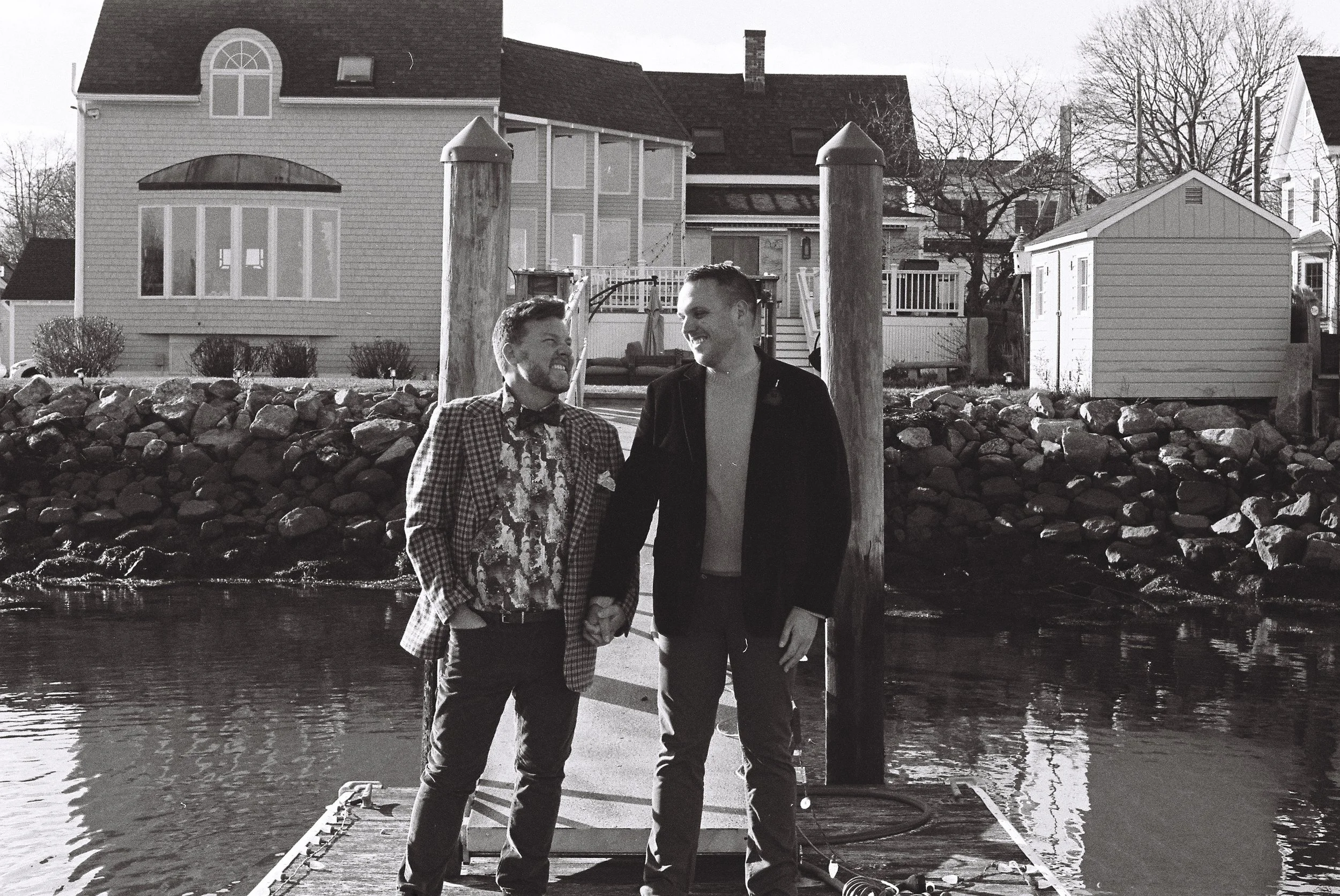 Two men standing on a dock by the water, holding hands and smiling at each other, with houses and trees in the background.