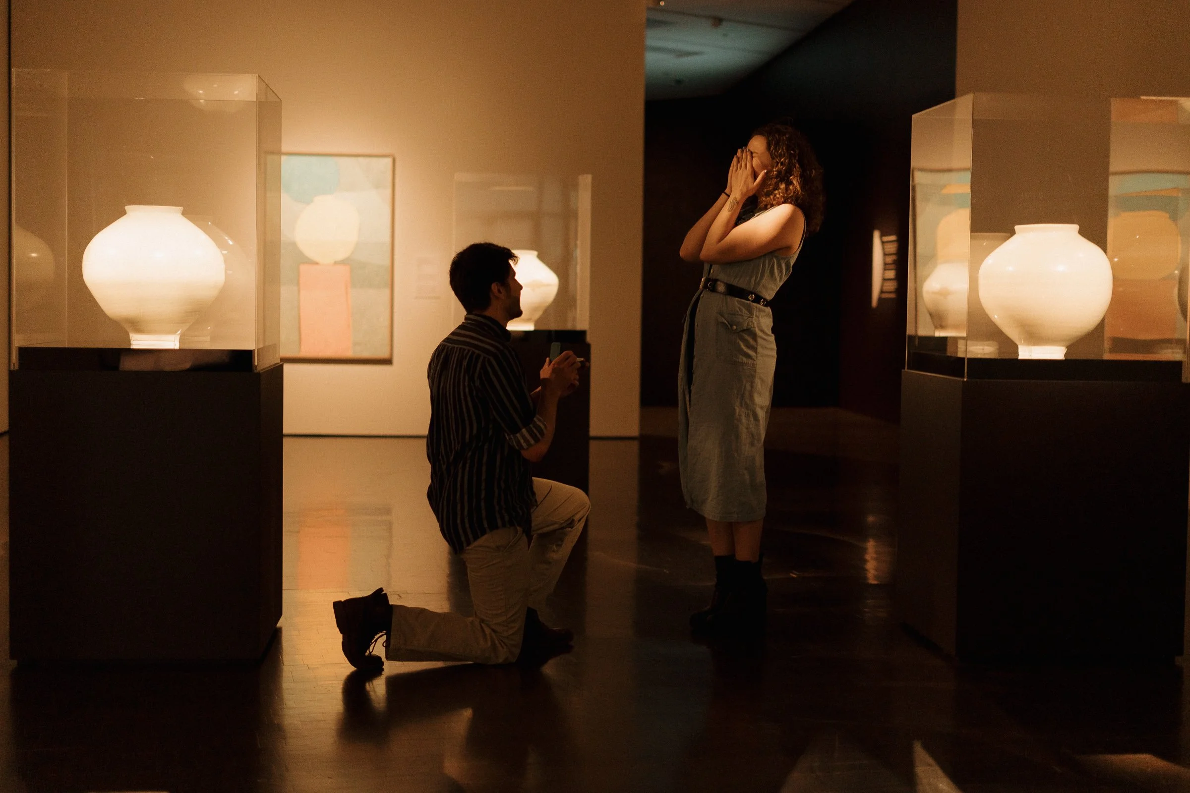 A man kneeling on one knee proposing to a woman in an art gallery, with framed artwork and illuminated vases in the background.
