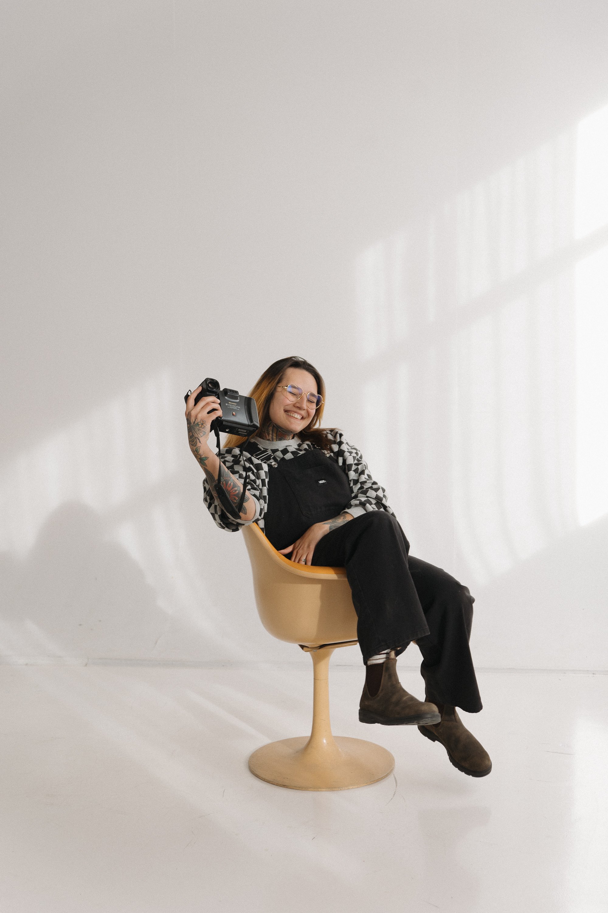 A woman sitting in a modern beige chair with a smile, holding a camera in her right hand, dressed in a checkered shirt, overalls, and brown boots, in a white room with sunlight streaming through large windows.