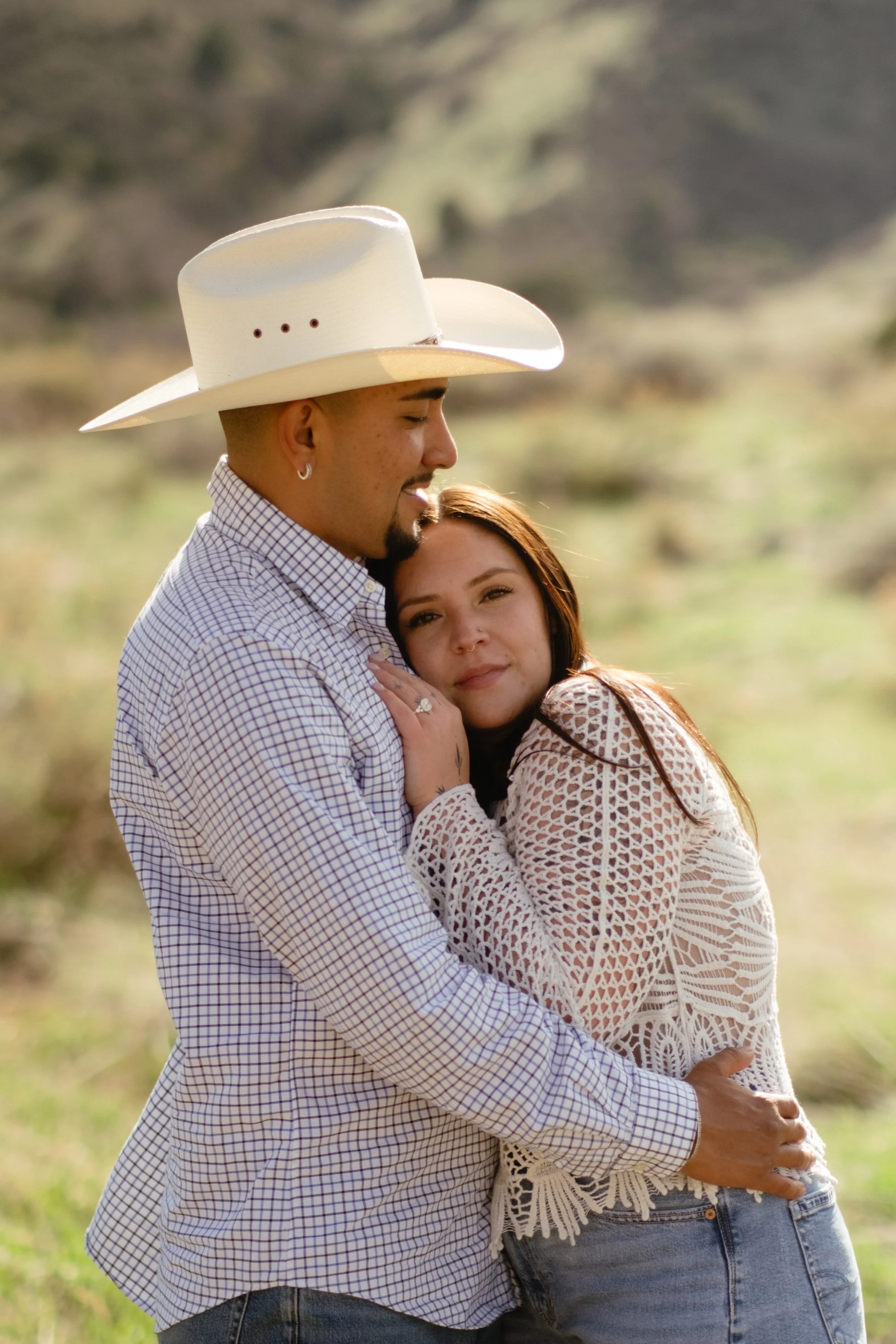 A couple standing outdoors, embracing, with a scenic blurred landscape in the background. The man wears a cowboy hat and a checkered shirt, and the woman has long hair and wears a white crochet top.