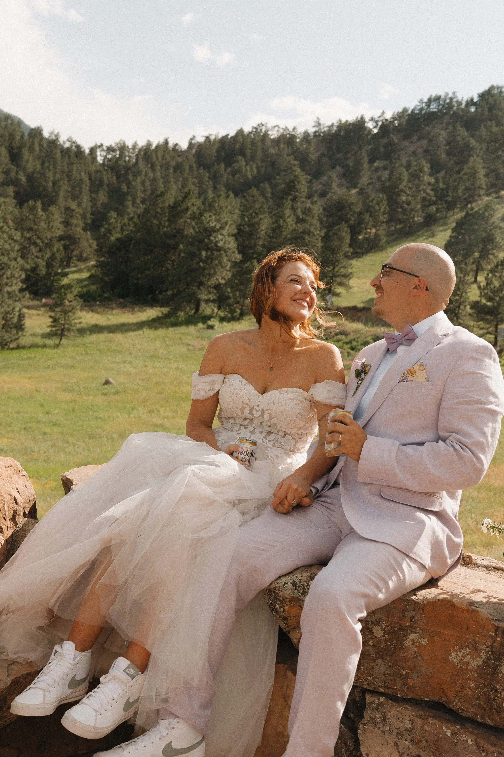 A woman in a white wedding dress and a man in a light-colored suit sit on a rock outdoors in a scenic mountainous area, smiling and holding cans of beer.