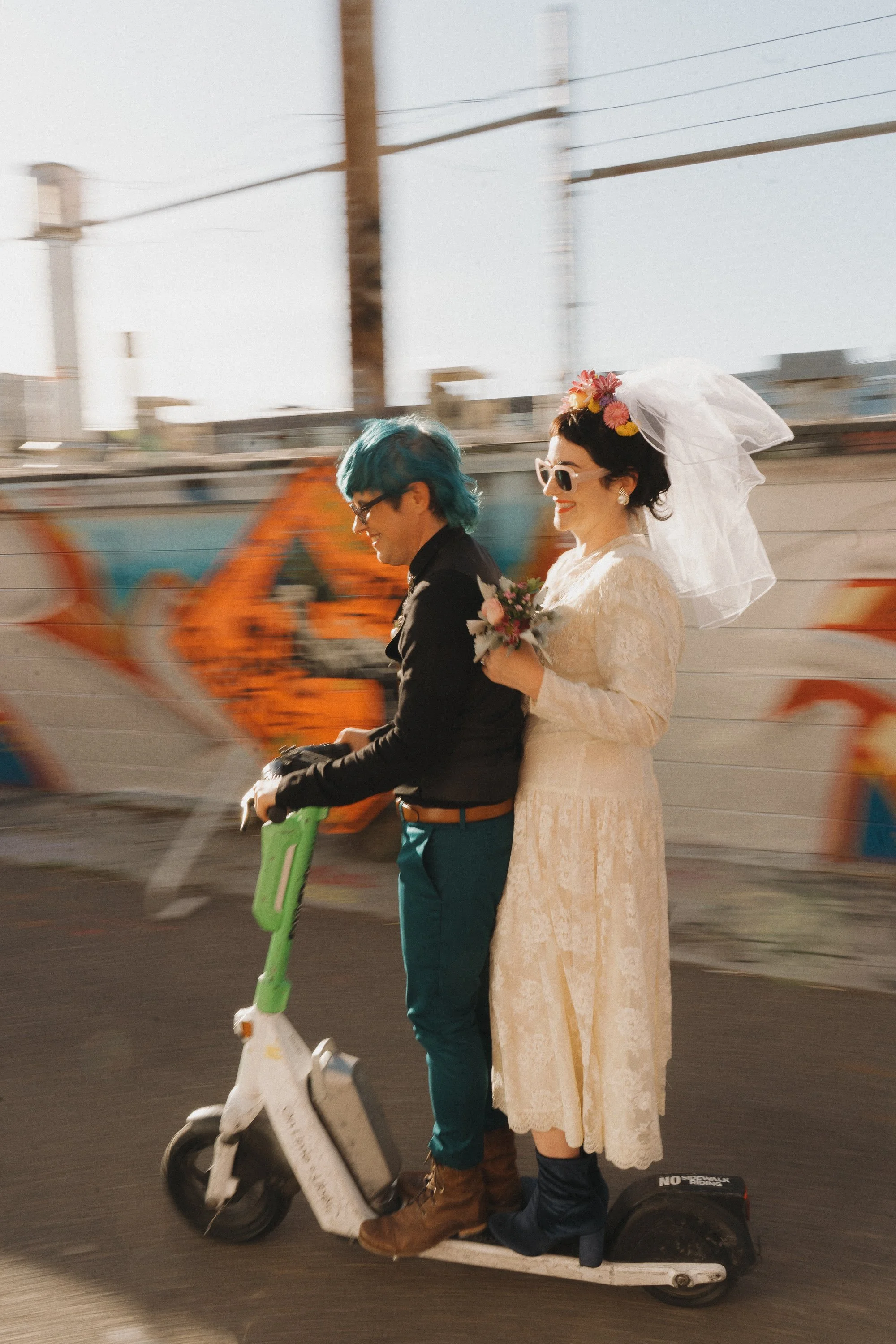 A bride and groom riding a scooter together against a graffiti-covered wall, both smiling.