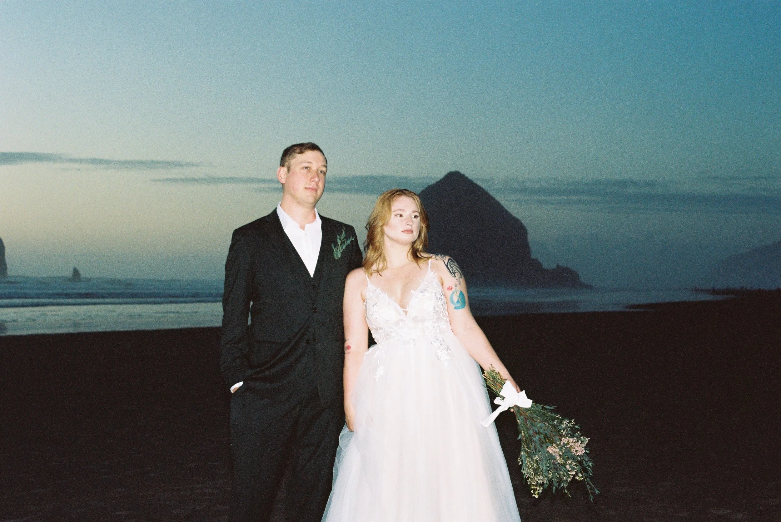 A bride and groom standing on a beach at sunset with cliffs in the background.