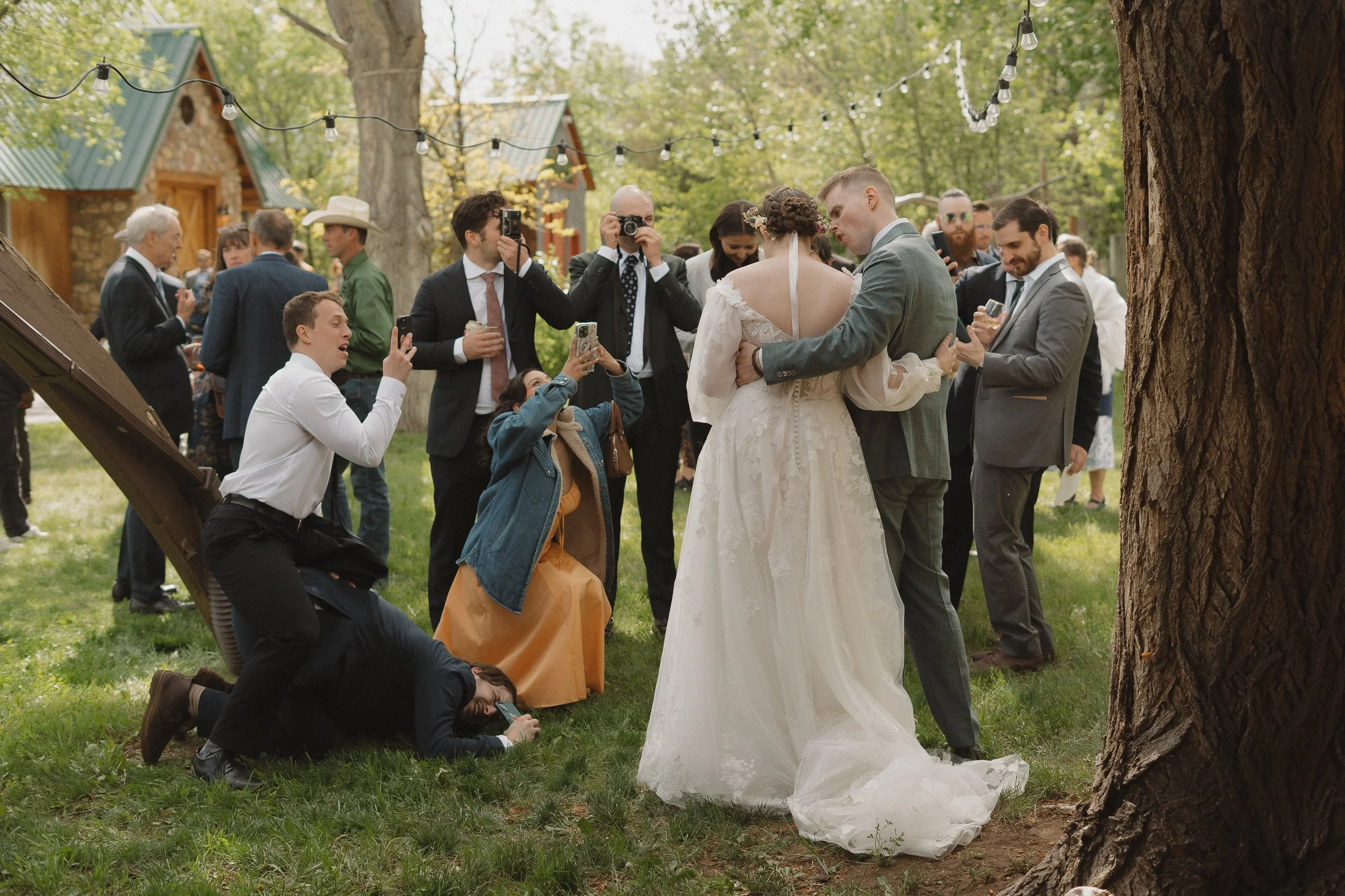 Bride and groom embracing at their wedding, surrounded by friends and family taking photos in an outdoor setting with trees and string lights.