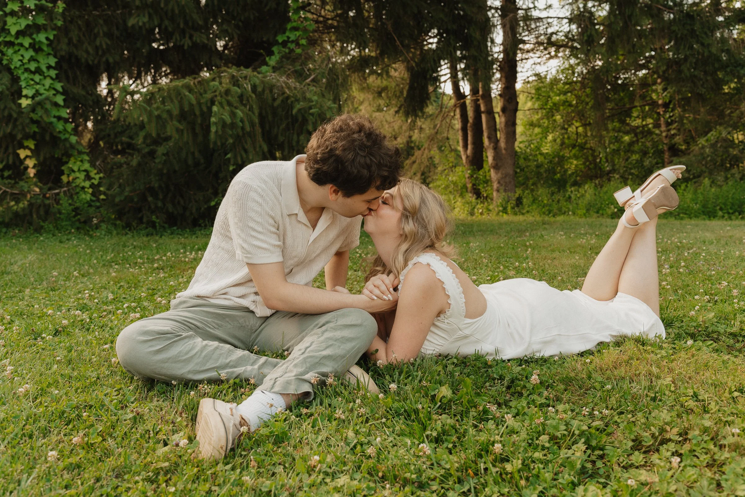 A couple is sharing a kiss outdoors on a grassy field surrounded by trees. The man is sitting cross-legged in a light-colored shirt and pants, while the woman is lying on her stomach in a white dress and sandals, with her legs bent at the knees.