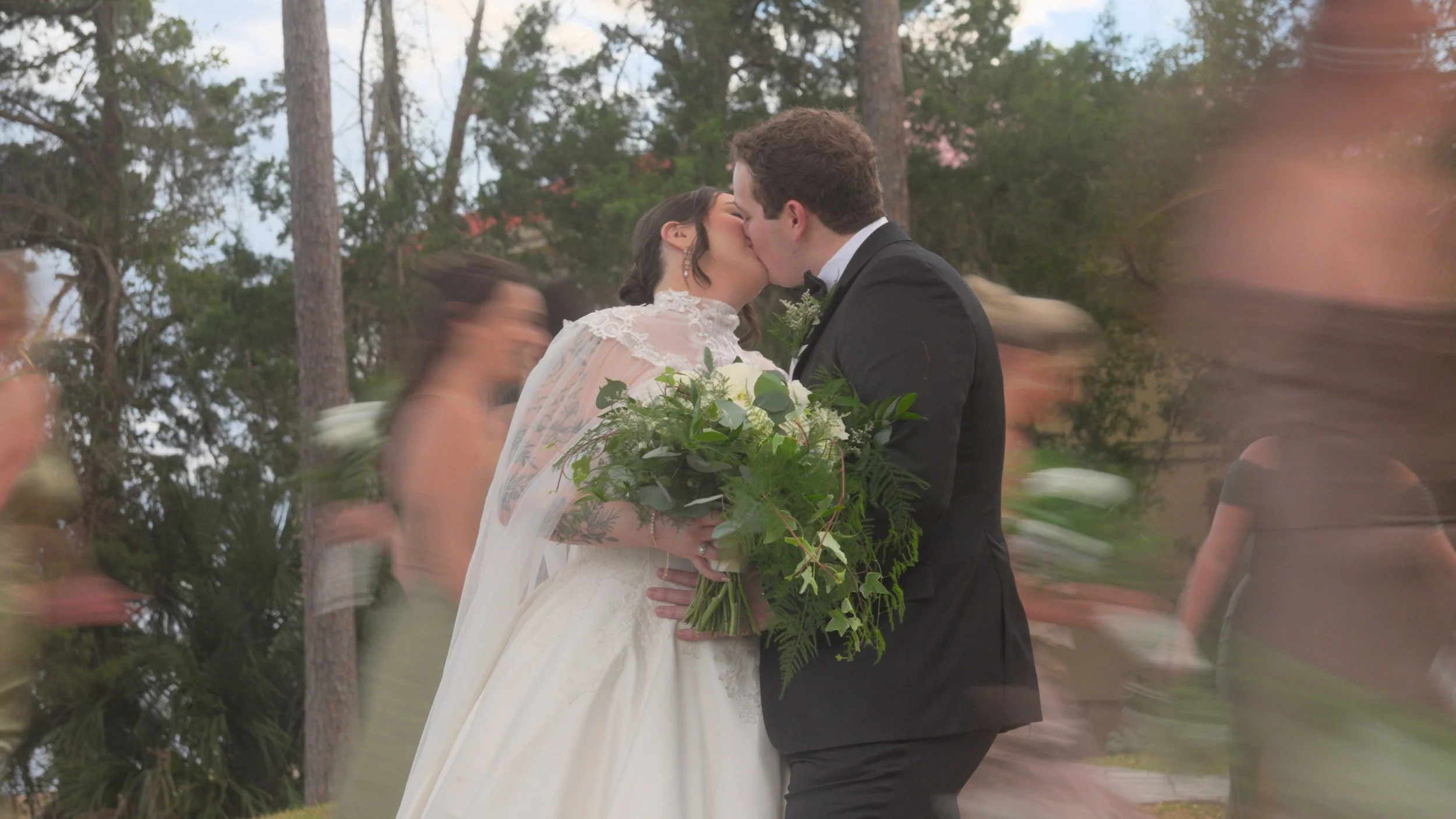 A bride and groom sharing a kiss during their wedding ceremony outdoors, with blurred guests in the background.
