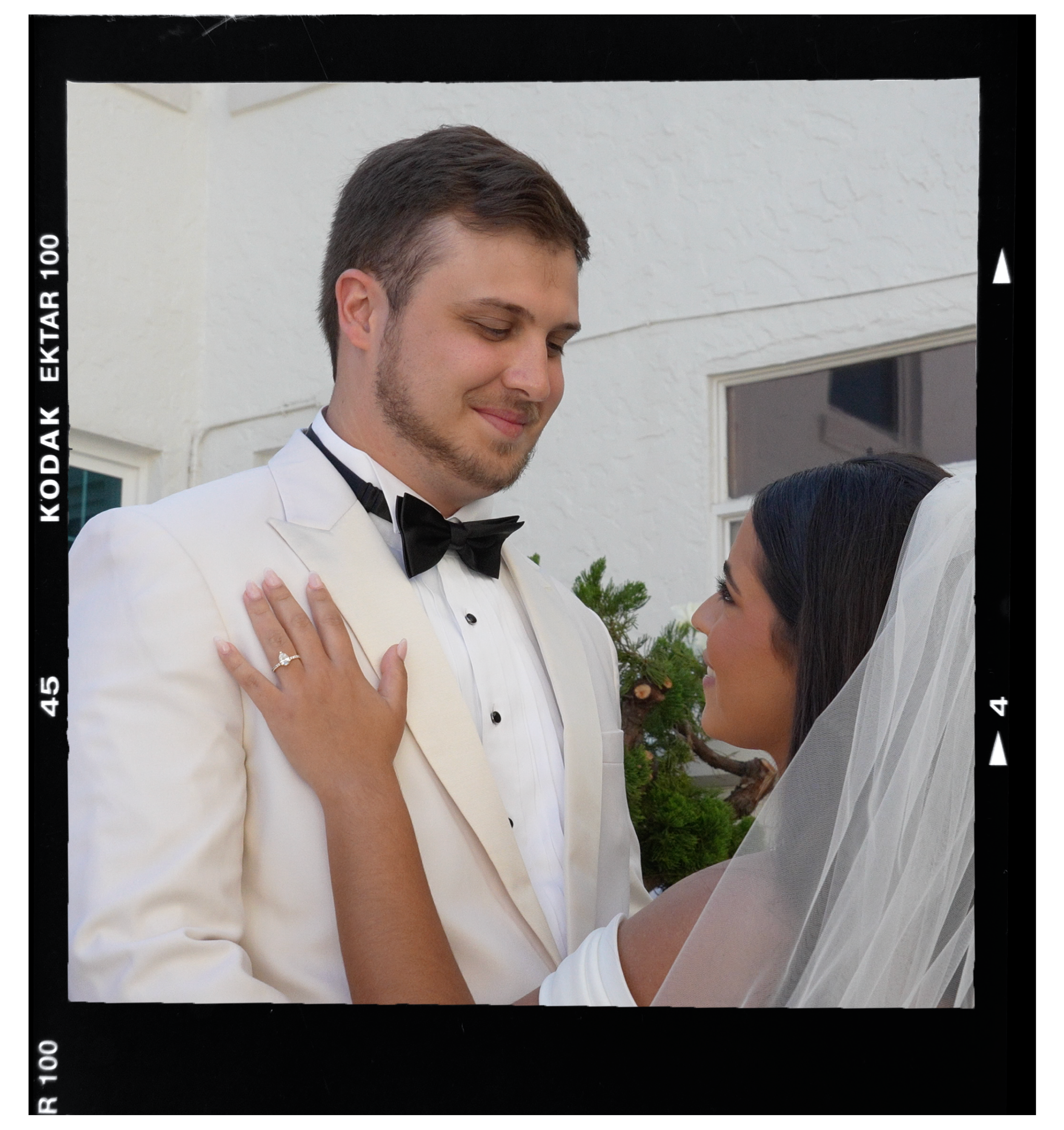 A bride and groom in wedding attire sharing an intimate moment, smiling at each other outdoors.