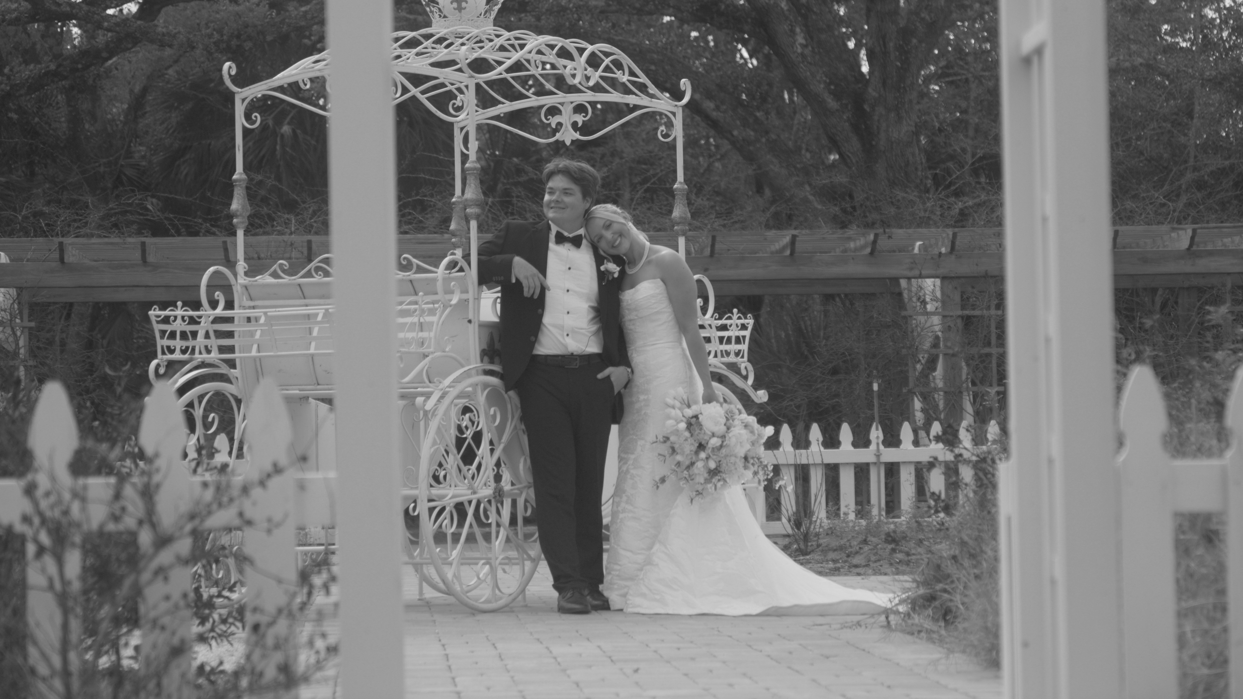 A bride and groom posing in front of a vintage carriage, close to a white fence with trees in the background, on their wedding day.