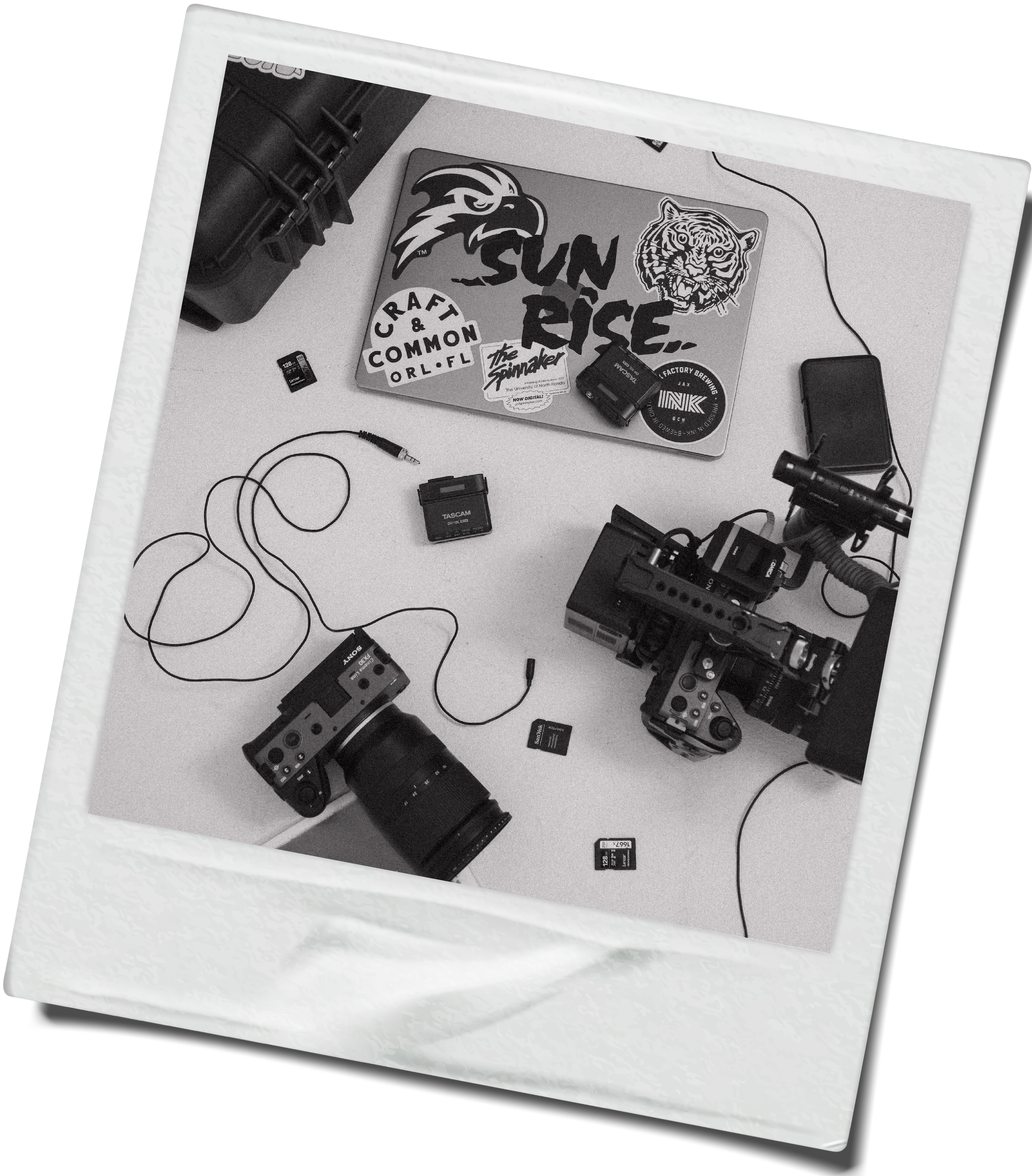 Arrangement of filmmaking equipment including cameras, memory cards, and cables on a white surface, with a laptop decorated with stickers.