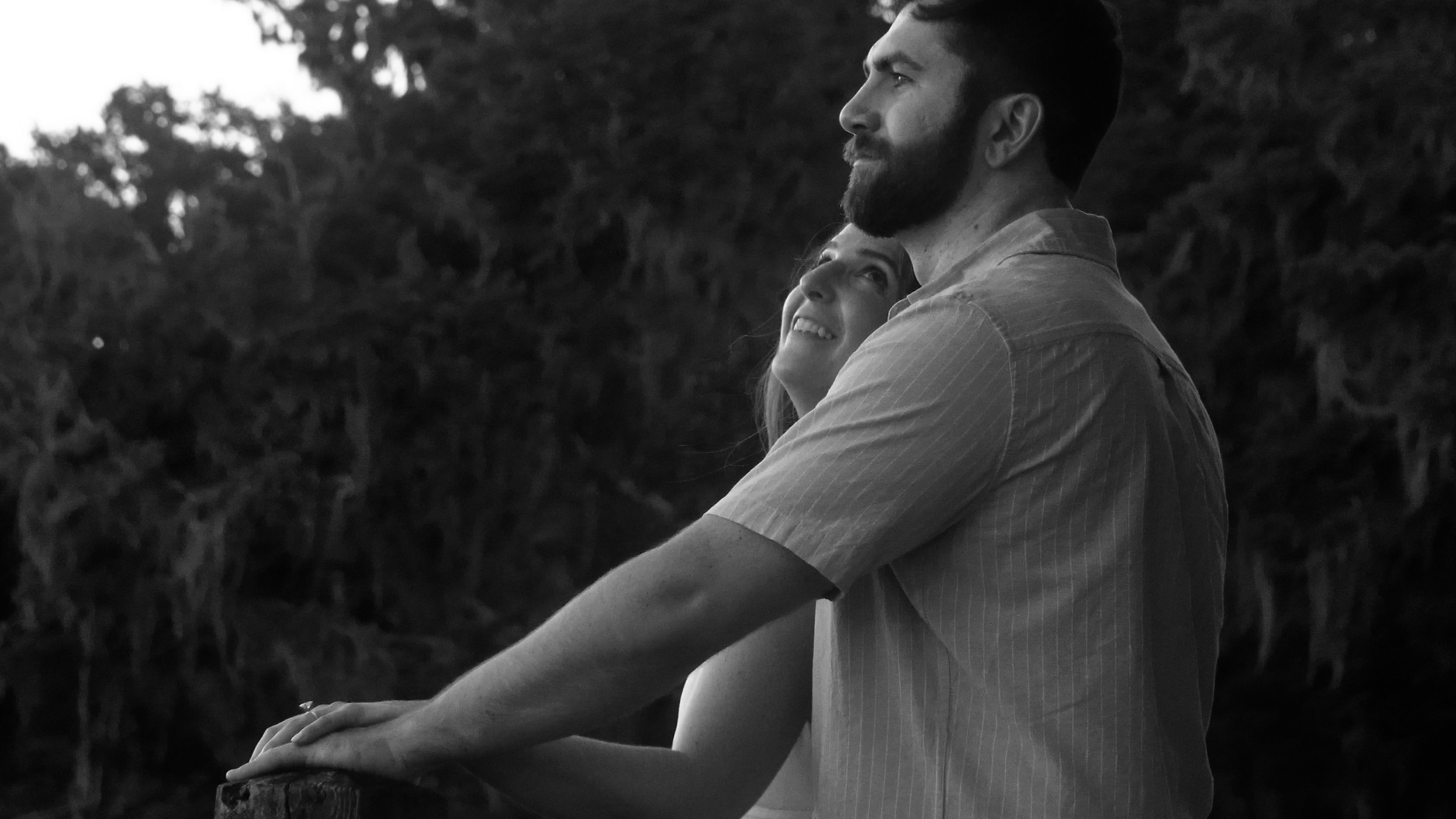 A couple leaning on a railing outdoors, with trees in the background, embracing and looking up. The photo is in black and white.