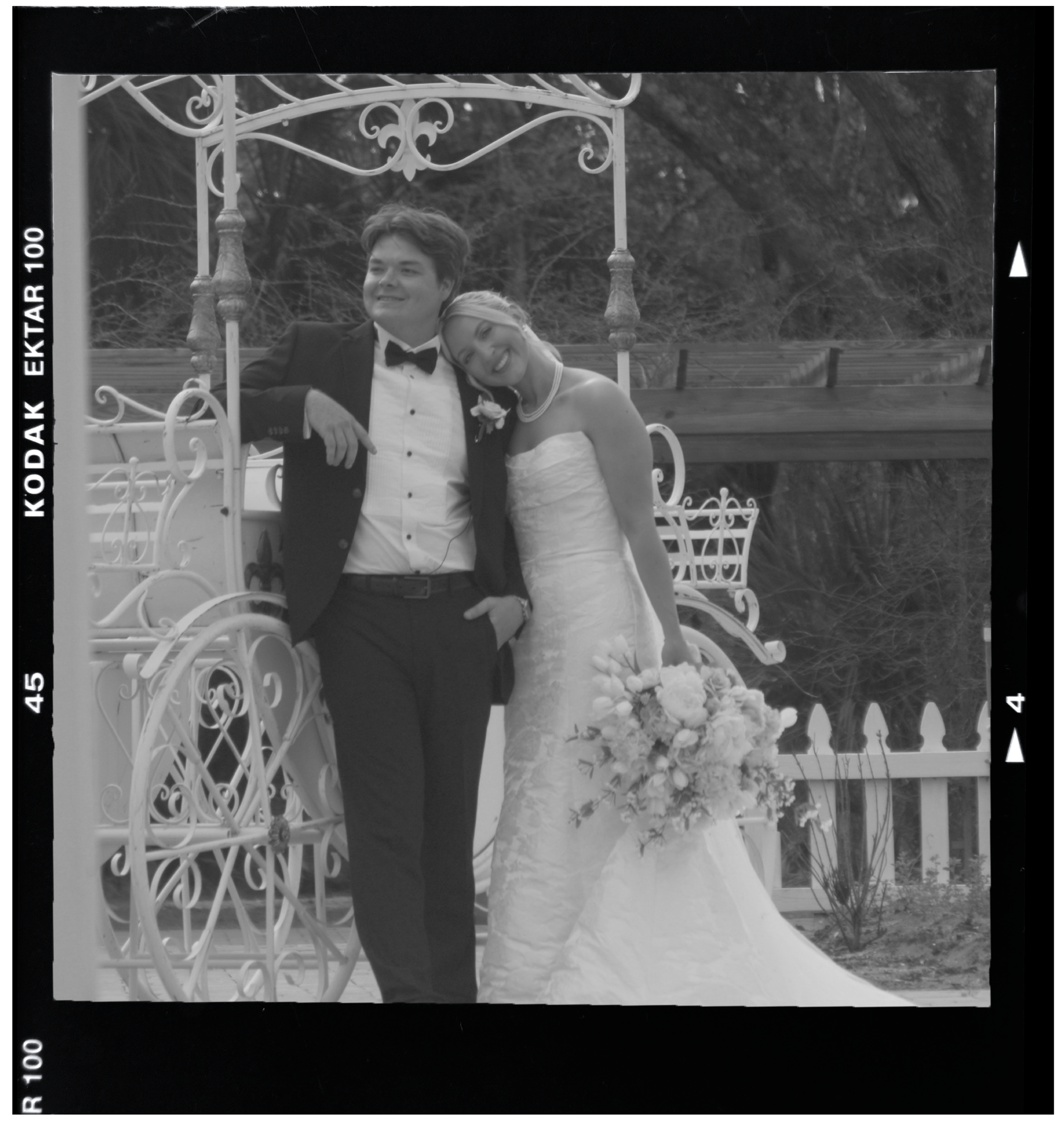 Black and white photo of a bride and groom standing in front of a decorative carriage, with the bride leaning her head on the groom's shoulder. The bride is holding a large bouquet of flowers, and they are smiling.