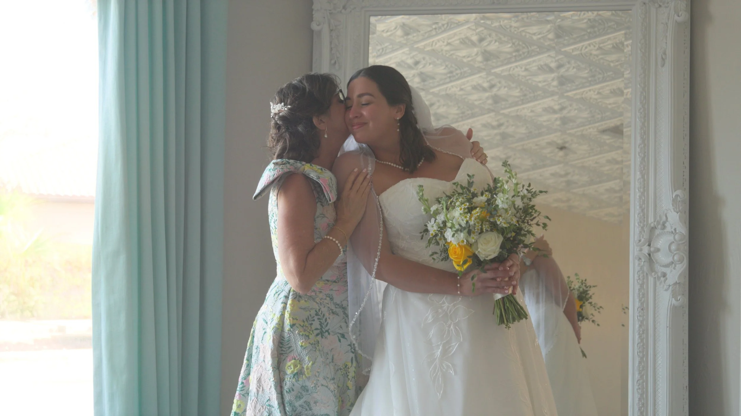 A bride in a white wedding gown with a bouquet of yellow and white flowers embraces and kisses a woman in a floral dress, both smiling, reflected in a mirror.