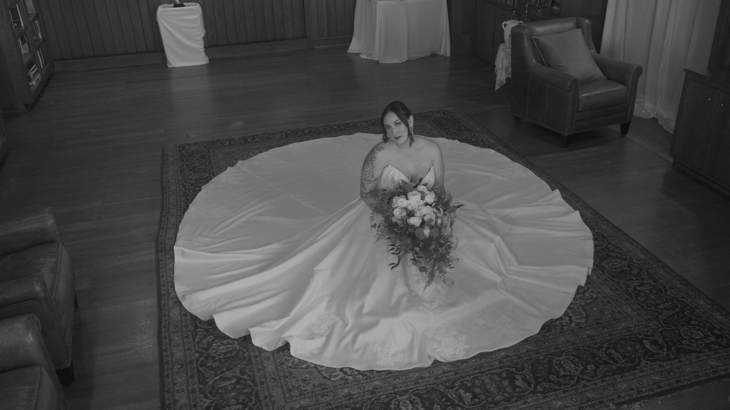 A woman in a wedding dress sitting on the floor on a large rug, holding a bouquet of flowers, in a room with wooden walls and furniture.