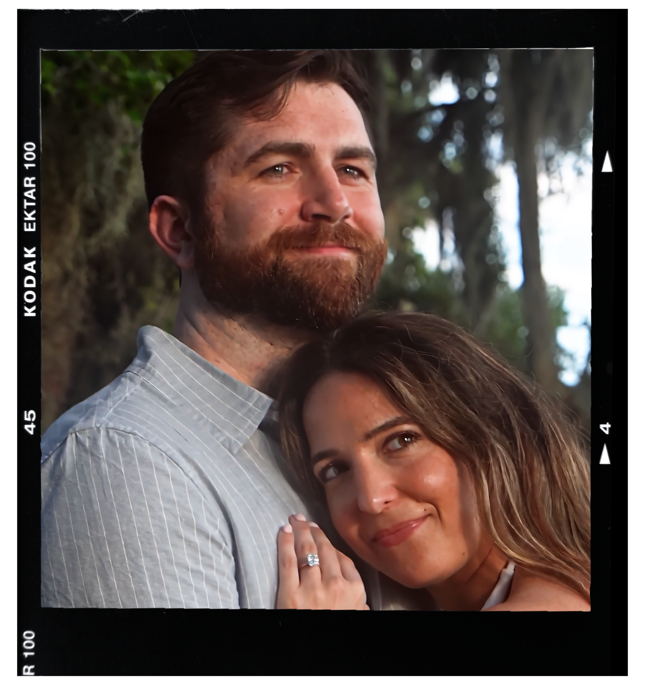 A man with a beard and a woman leaning on his shoulder, both smiling, in an outdoor setting with trees and natural light.